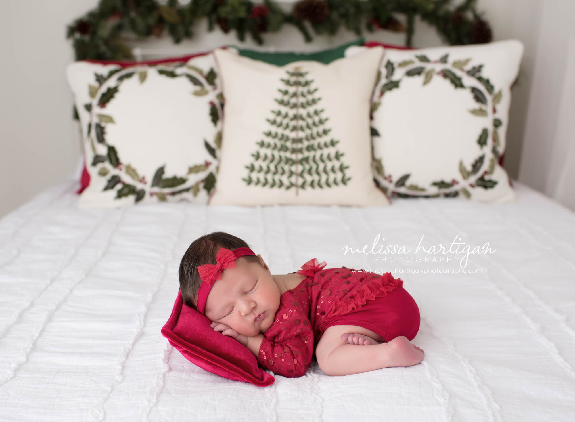 Melissa Hartigan Photography Connecticut Newborn Photographer trumbull Ct baby Fairfield county Baby girl Christmas theme red outfit with red headband on red pillow on bed with Christmas pillows sleeping Ct mini newborn session