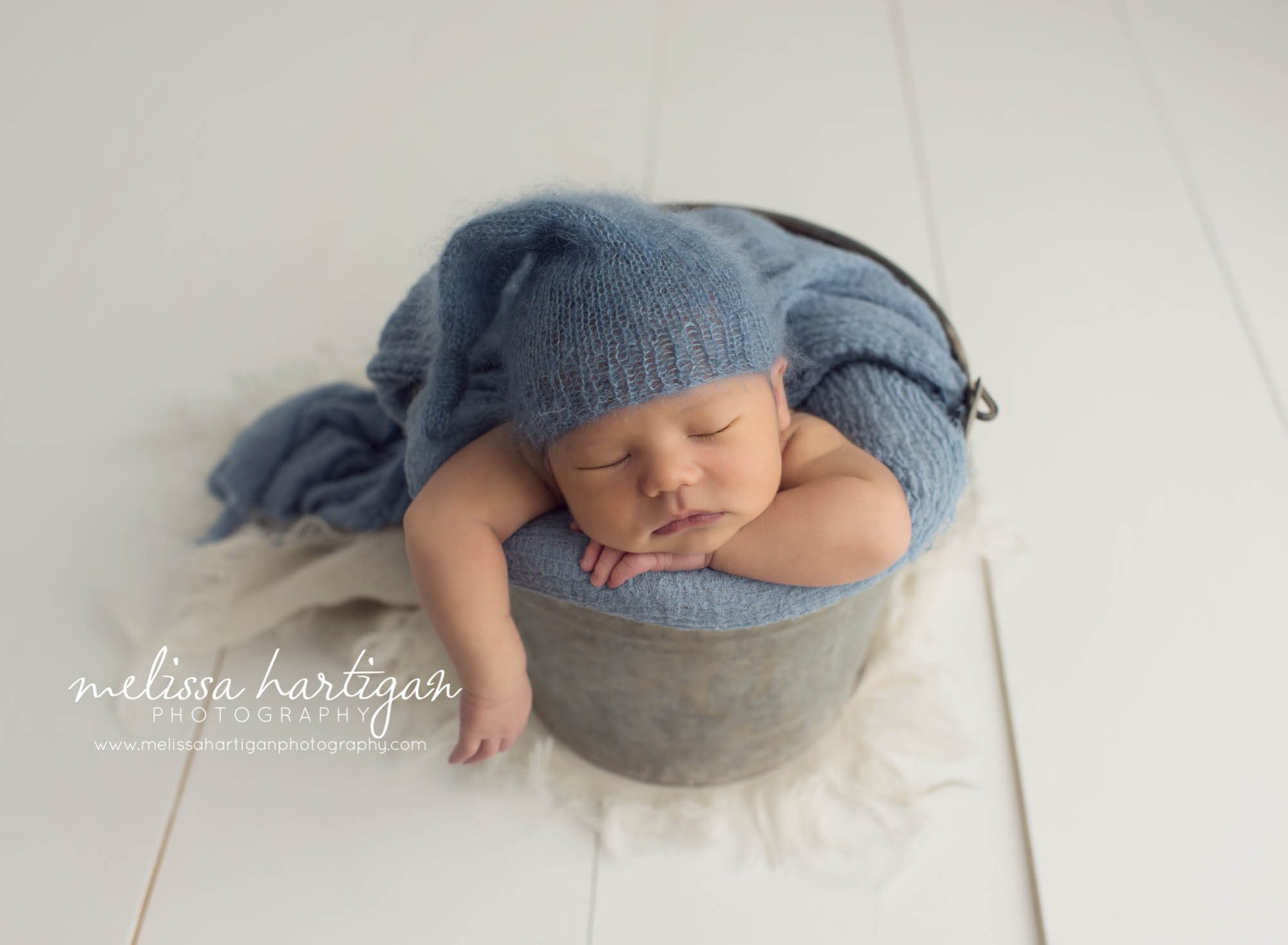 Melissa Hartigan Photography Maternity and Newborn Connecticut Photographer Lucas Newborn Session Baby boy sleeping in metal bucket with blue knit blanket and matching blue knit hat with arm sticking out