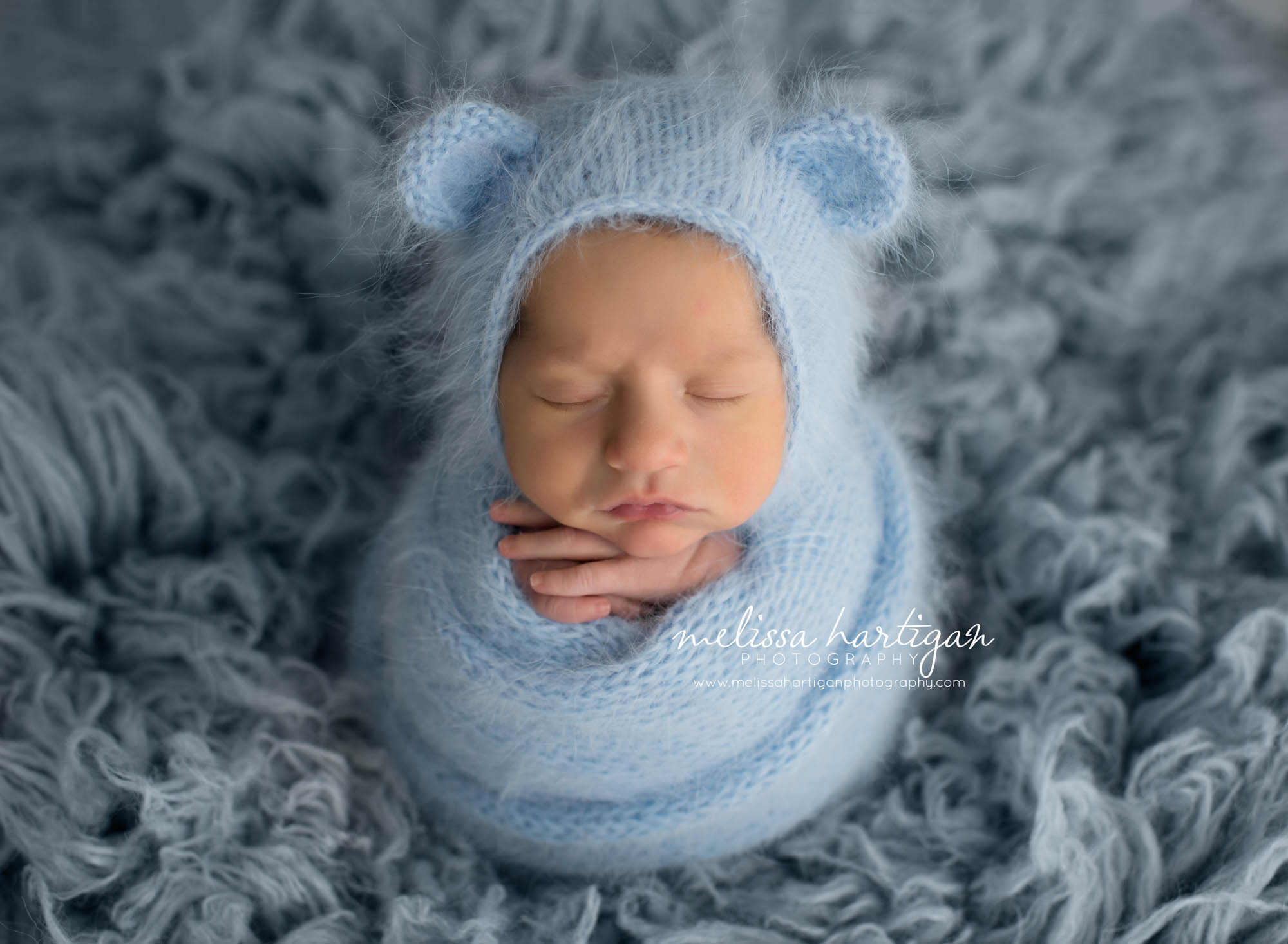 Melissa Hartigan Photography Maternity and Newborn Connecticut Photographer Brayden Mini Newborn Session Baby boy wrapped in blue knit blanket sleeping on a blue flokati hands sticking out wearing blue knit hat with ears