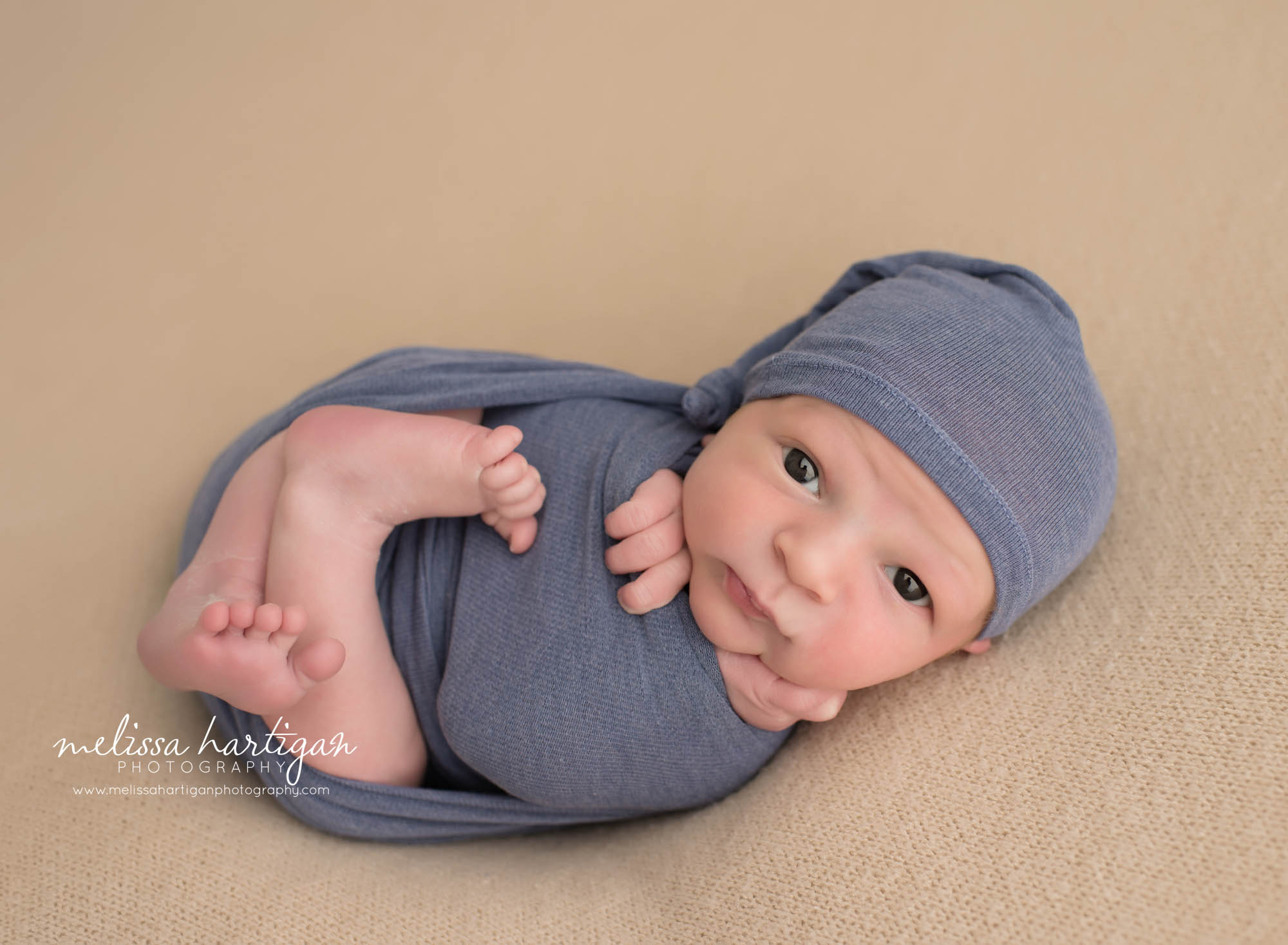 Melissa Hartigan Photography Newborn Photographer Connecticut baby boy wearing blue knit hat and matching wrap laying on tan blanket awake