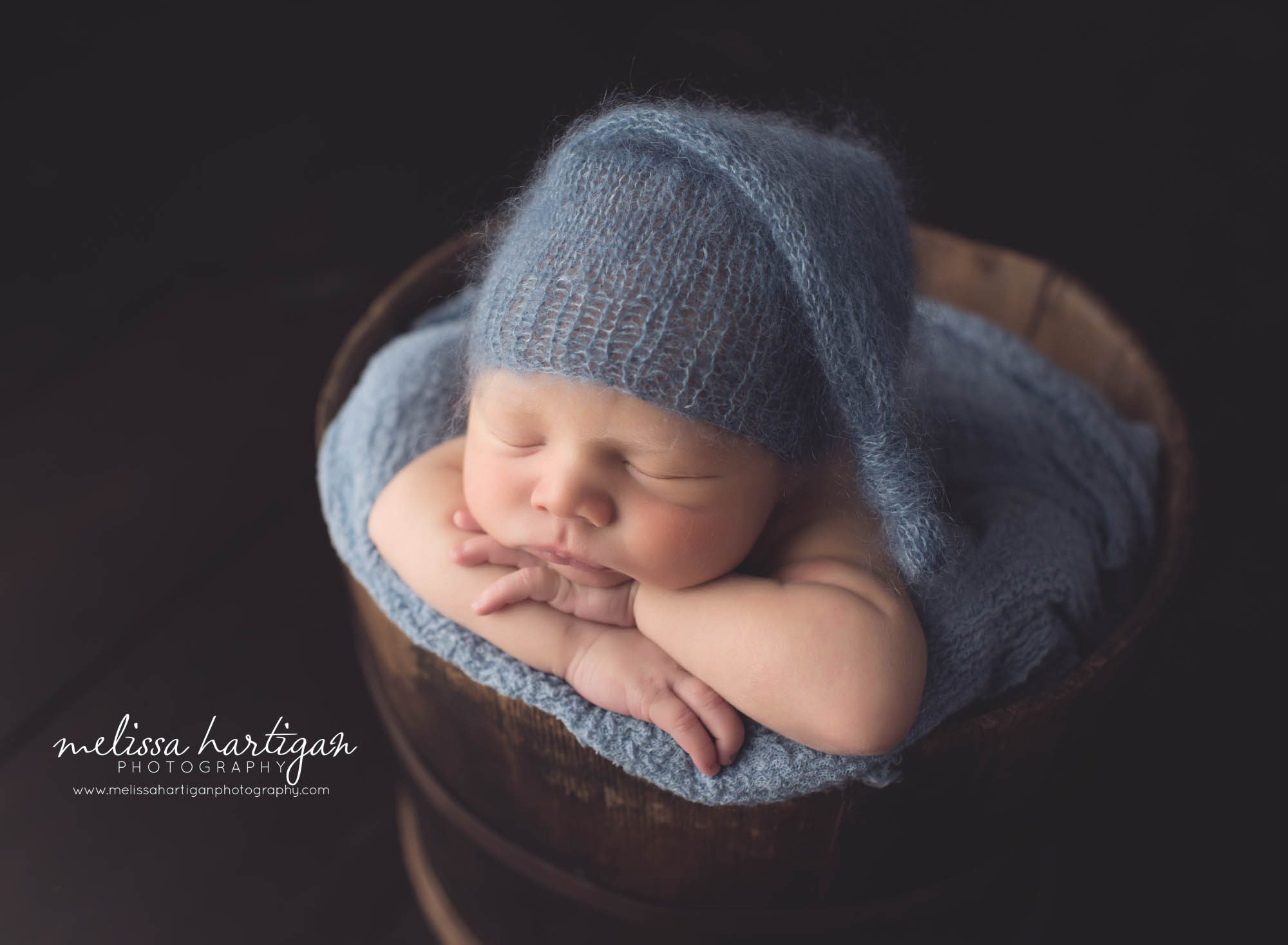 Melissa Hartigan Photography CT Newborn Photographer Braeden Newborn Session baby boy sleeping in wooden bucket wearing blue knit hat with blue knit blanket head on crossed arms
