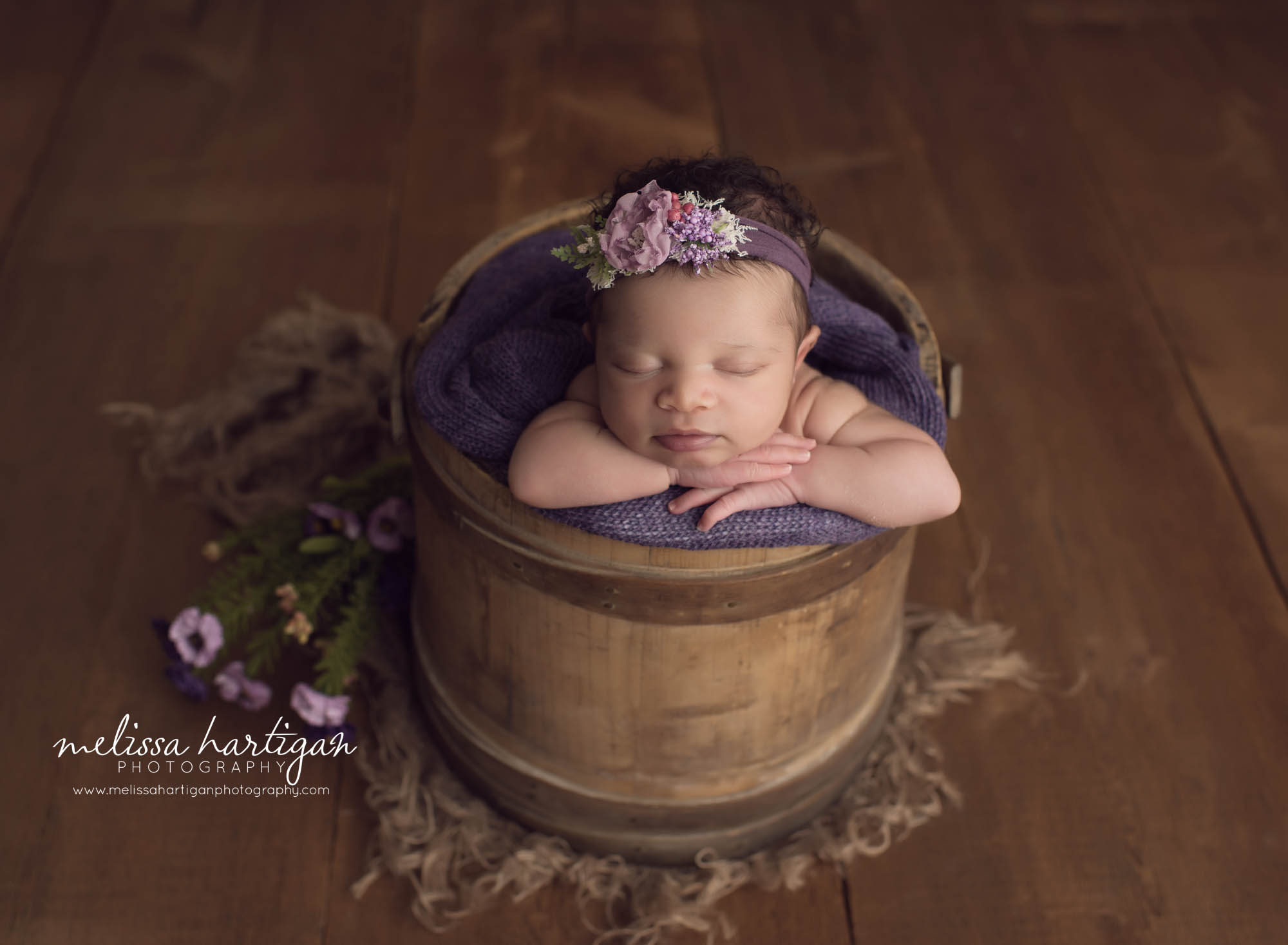 Baby girl posed chin on hands in rustic bucket with purple tones and flowers newborn photography session in CT studio