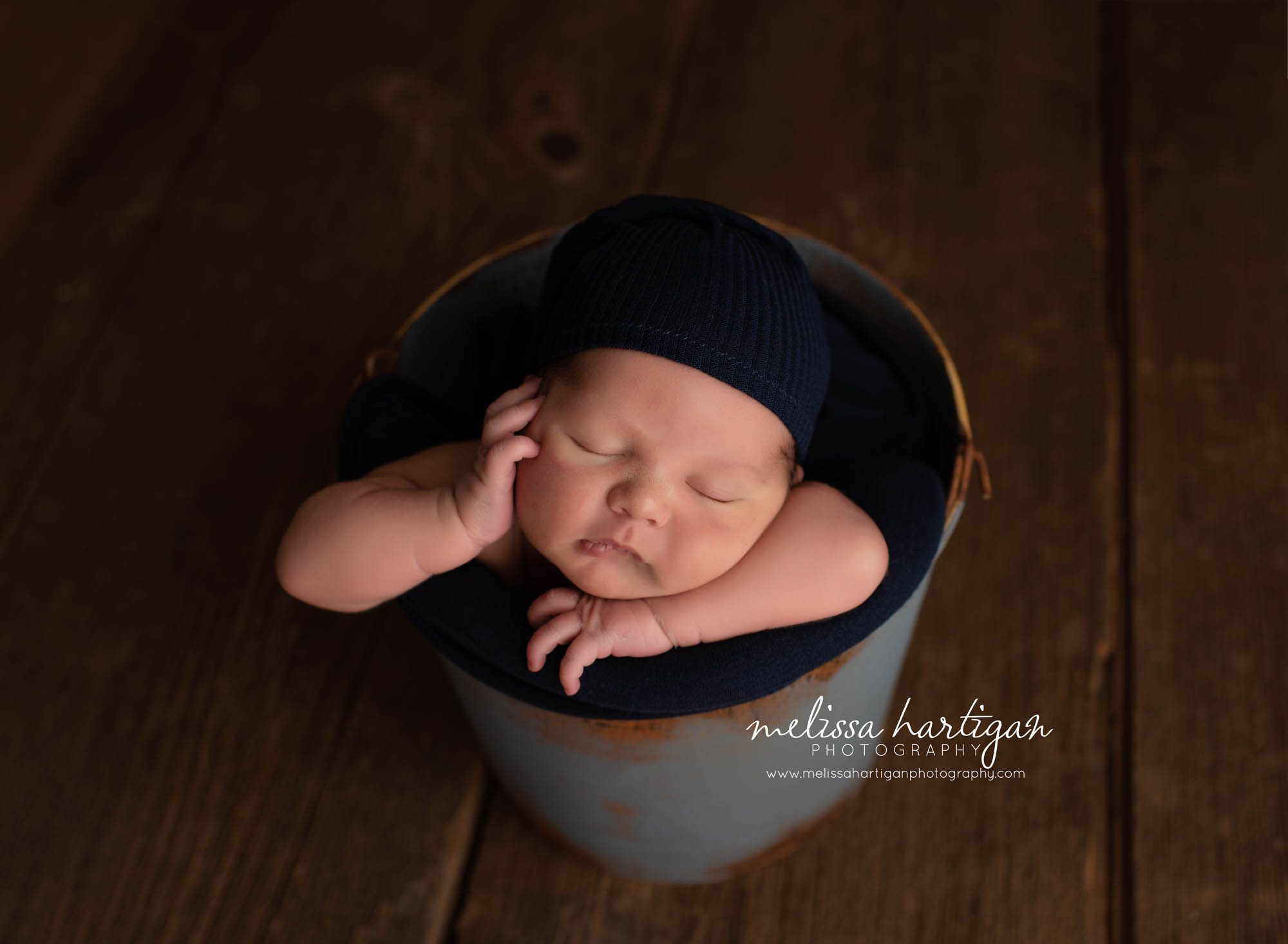 Baby boy posed in bucket with hand on cheek wearing navy blue hat
