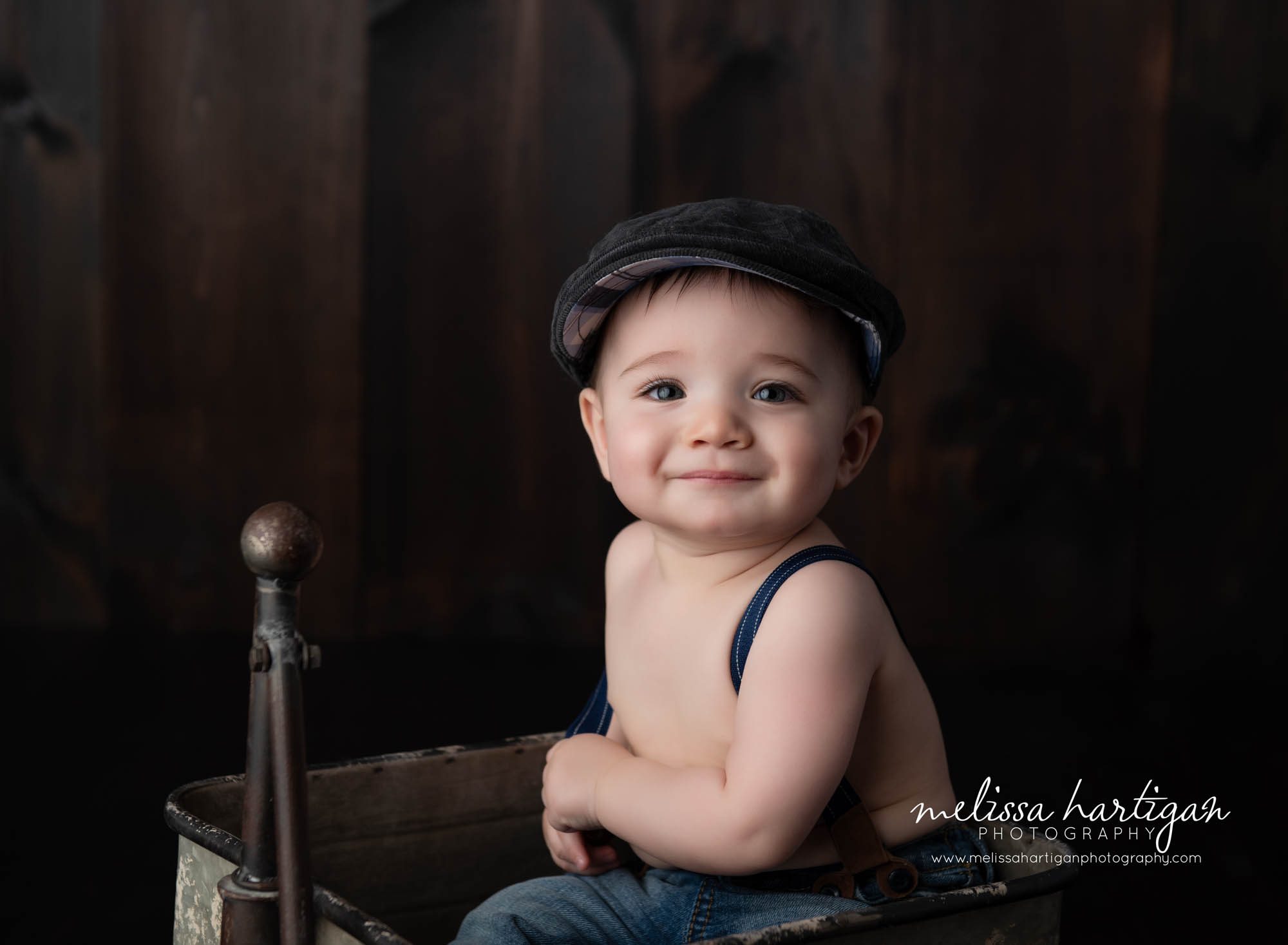 Baby boy looking at camera wearing jeans and suspenders sitting in old metal wagon CT baby milestone Photographer