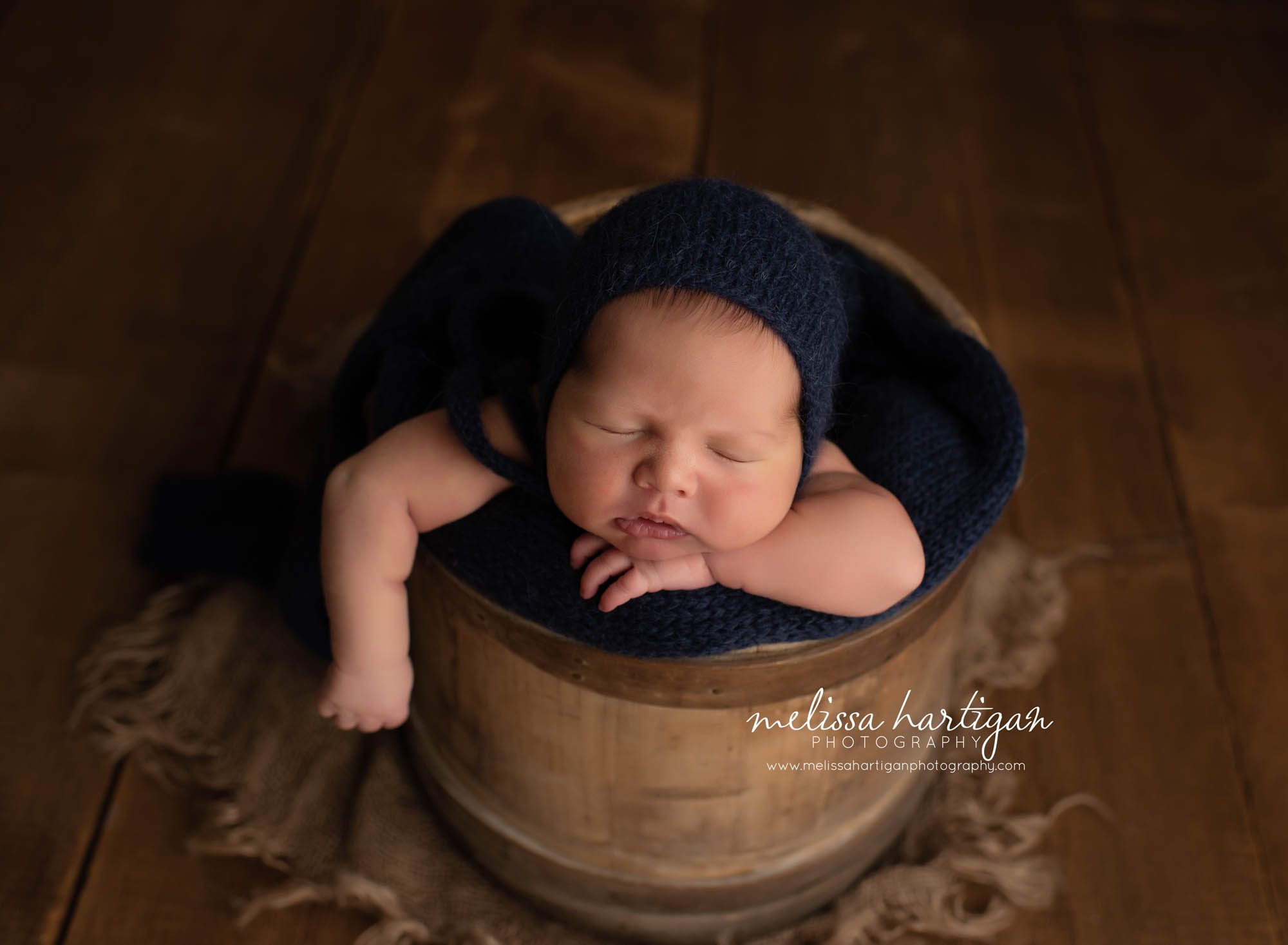 Newborn Baby boy wearing knitted navy blue hat posed in rustic bucket with arm hanging out