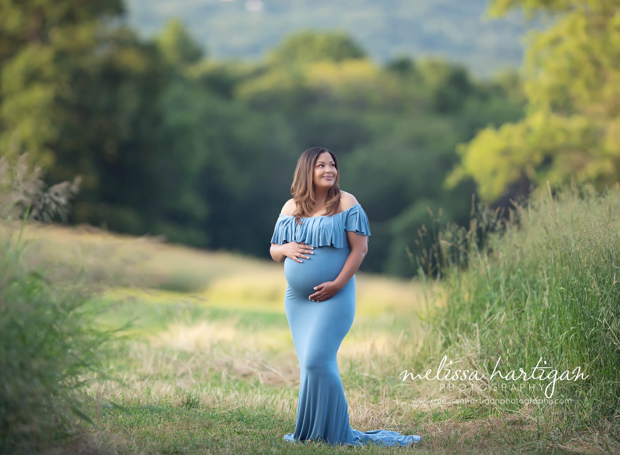 Expectant mom wearing long blue off the shoulder maternity dress standing on a hill with trees and grass