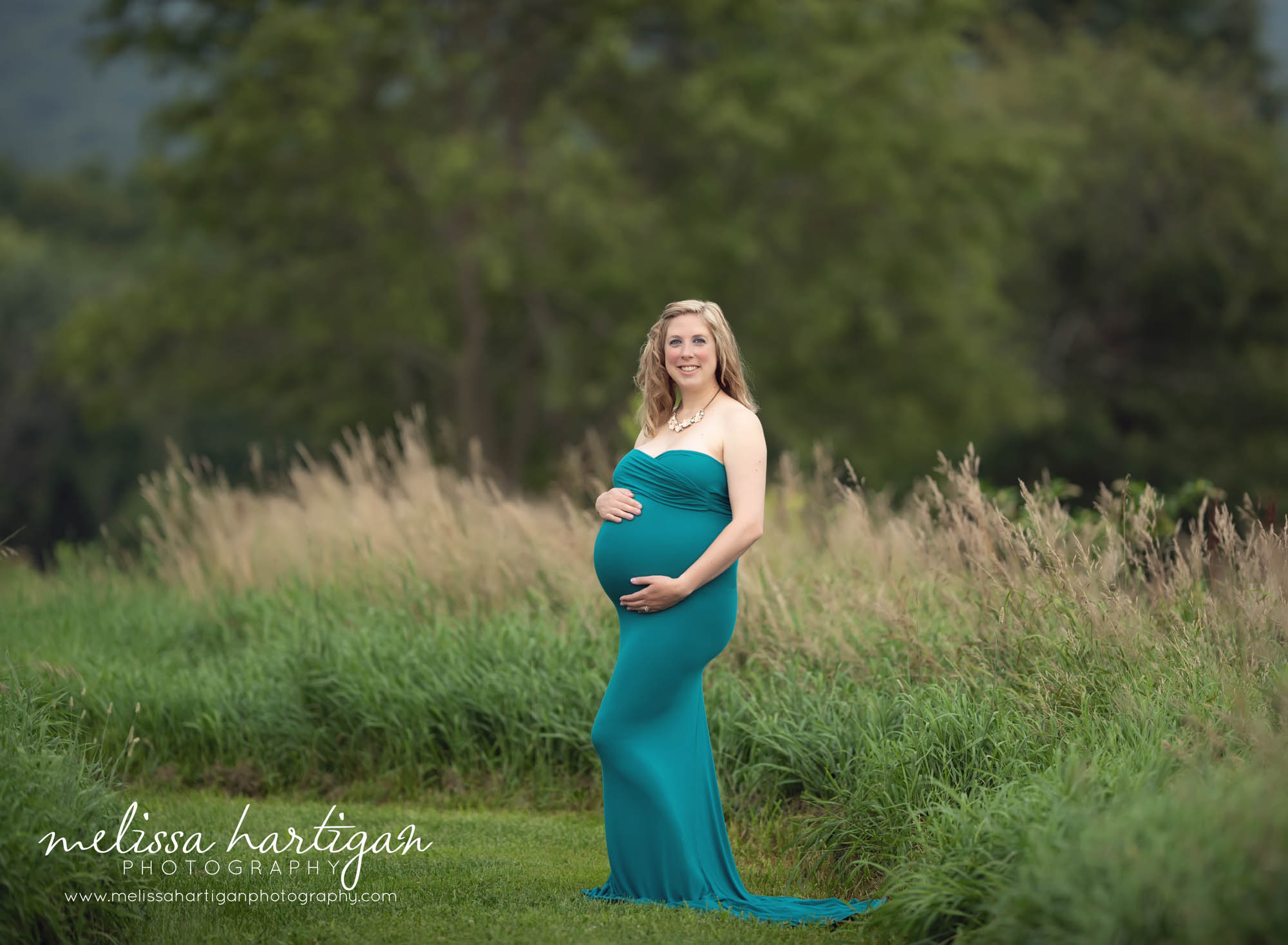 Expectant mother standing in grassy field area wearing peacock blue turquoise long fitted maternity dress sweetheart neckline maternity photographer columbia CT