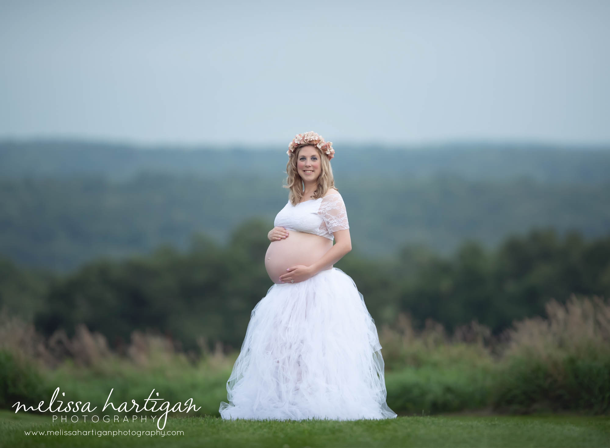 Pregnant mom standing on hill holding her baby bump wearing white tulle shirt and white lace crop top with sleeves maternity photography CT