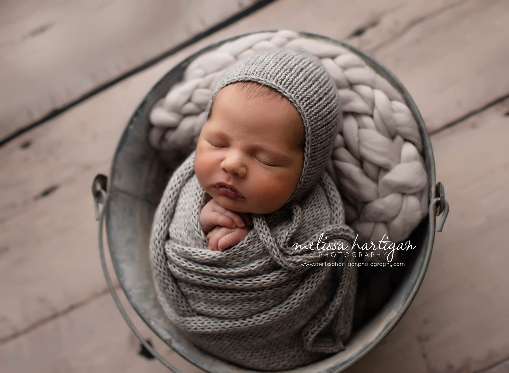 Baby boy wrapped in grey knitted wrap with grey knitted boy bonnet posed in metal bucket with light grey braided layer