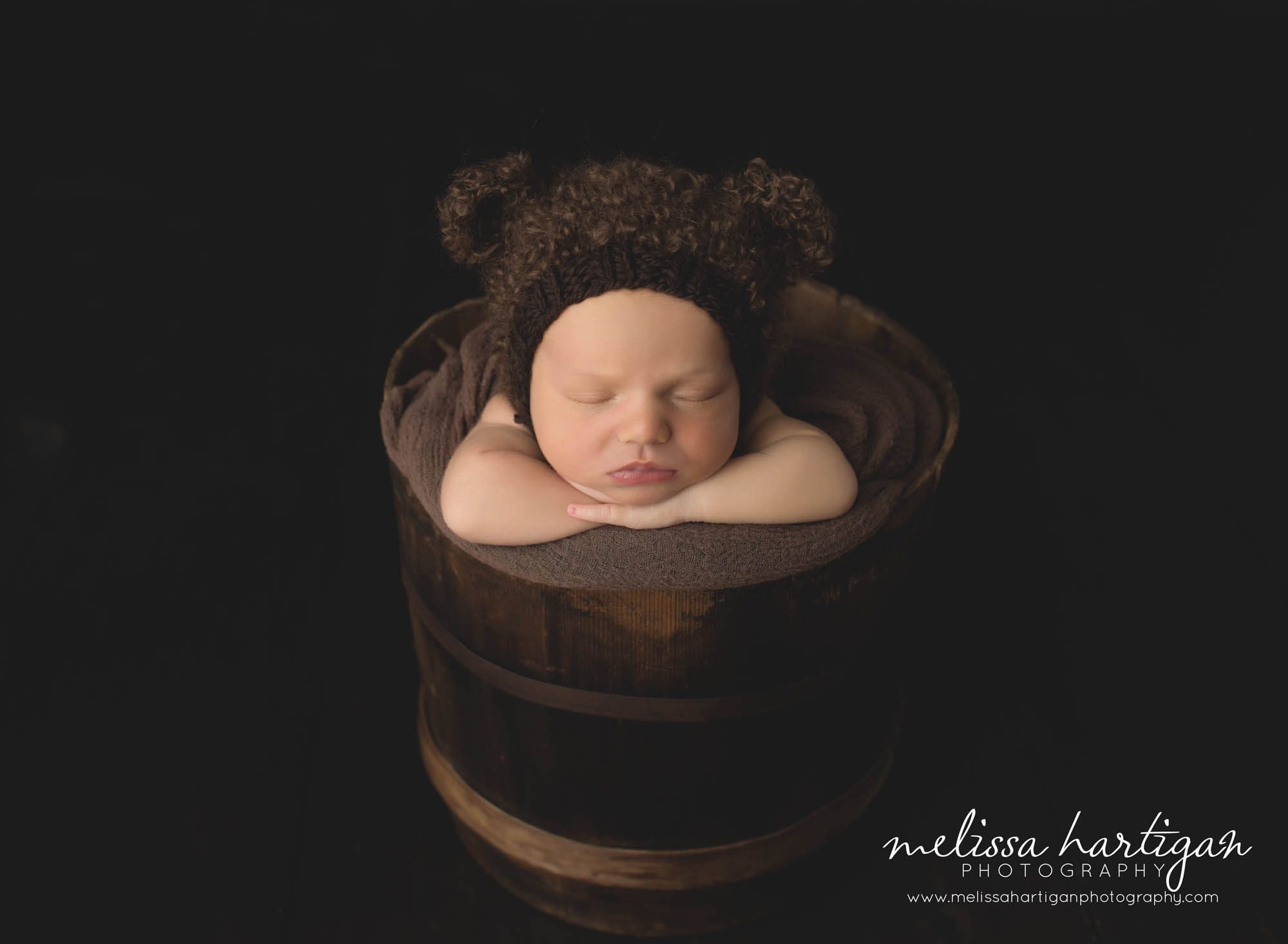 newborn boy posed in bucket wearing dark brown knitted bear bonnet