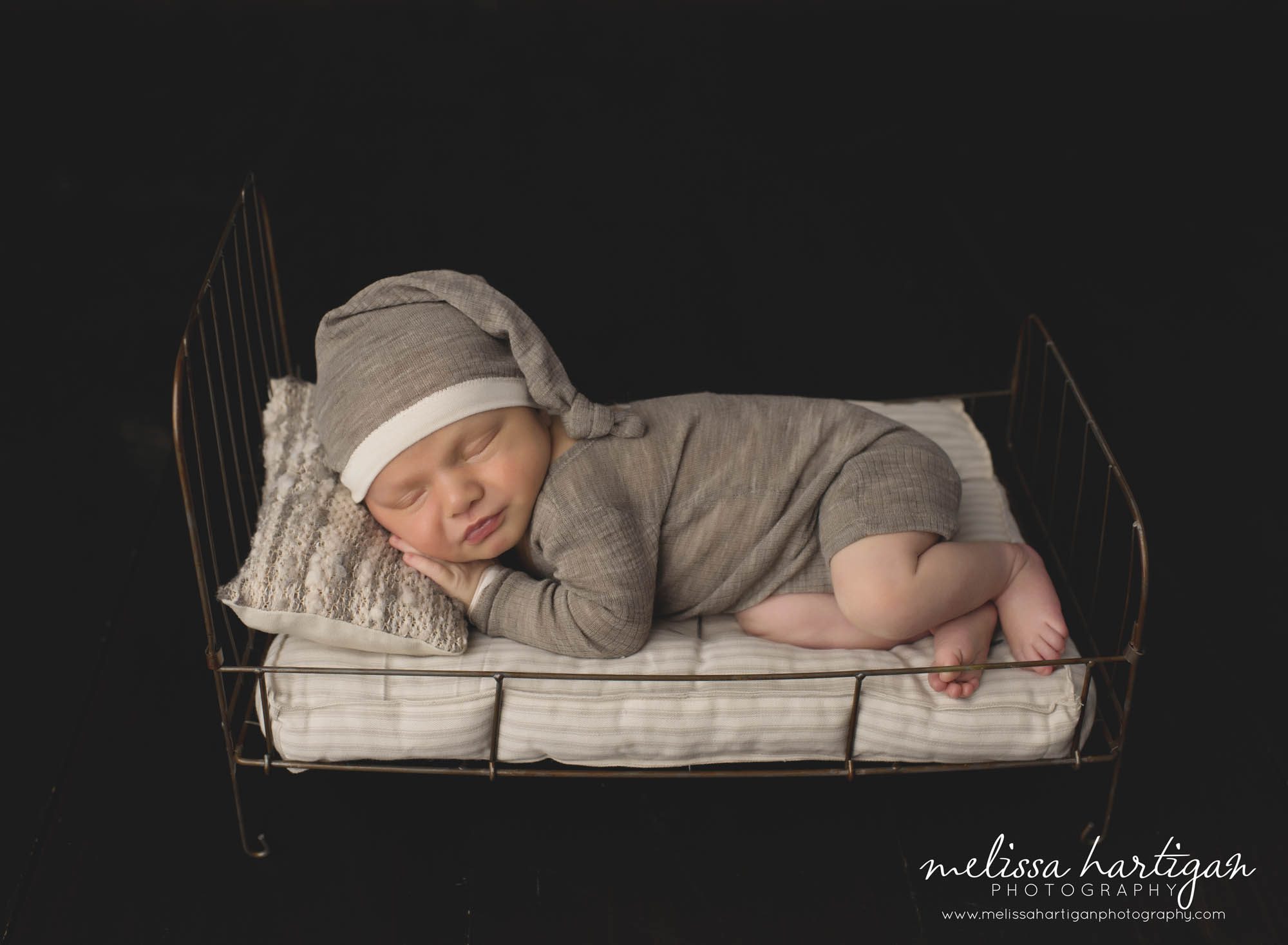 baby boy posed on metal prop bed wearing neutral colored sleepy hat and outfit