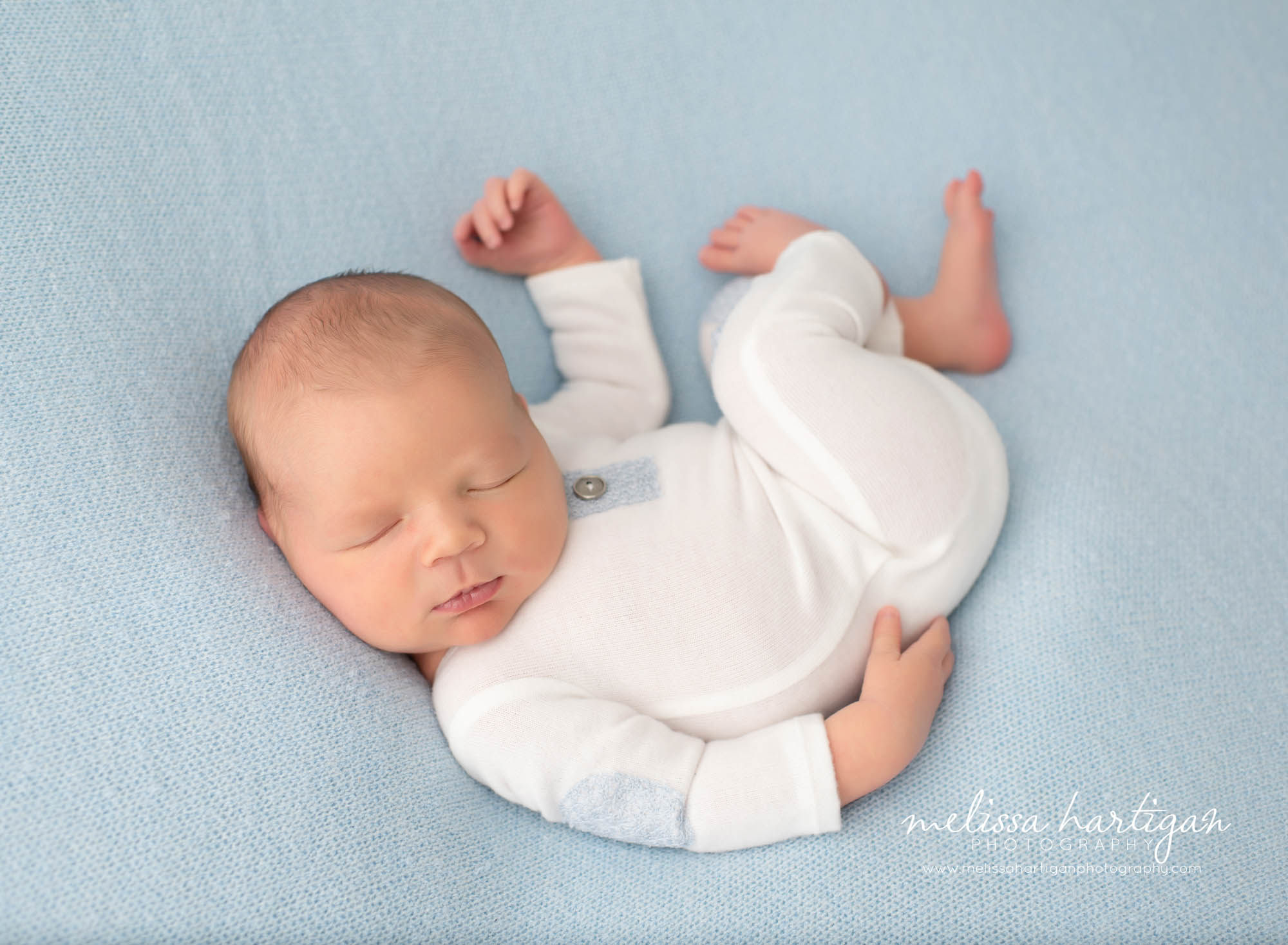 Baby boy wearing white and blue romper curled up on blue backdrop newborn photos CT