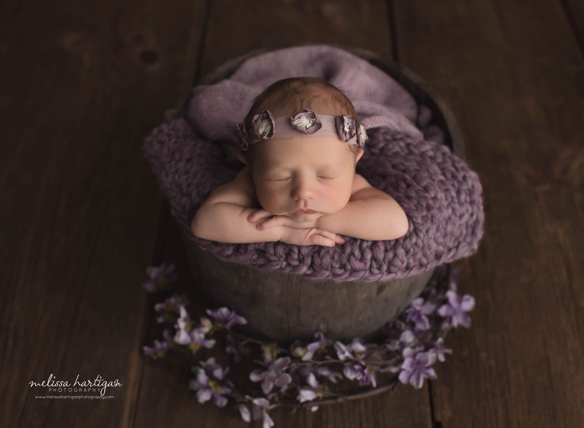 Baby girl posed in bucket with head resting on hands purple flowers placed around the bucket CT newborn Photographer