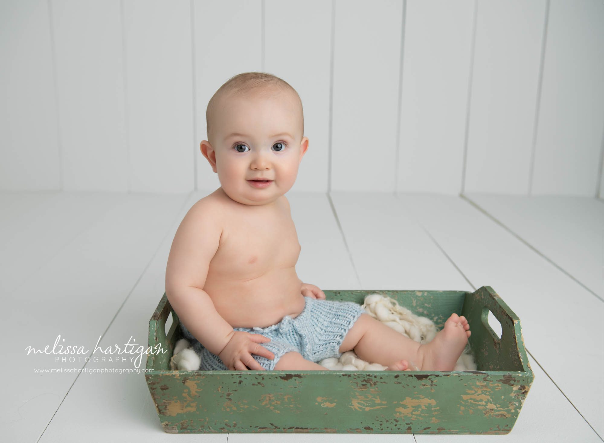 baby boy sitting in green wooden tray wearing light blue knitted shorts