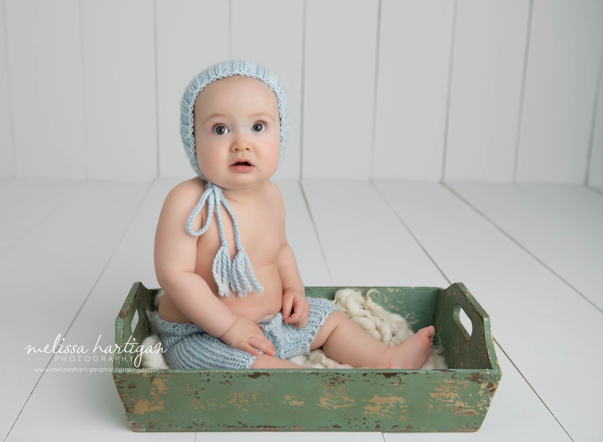 baby boy sitting in wooden green tray wearing light blue knitted bonnet