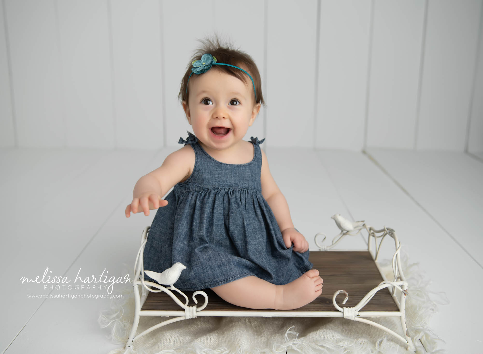 Baby girl sitting on vintage cream wire bed with birds wearing blue jean dress