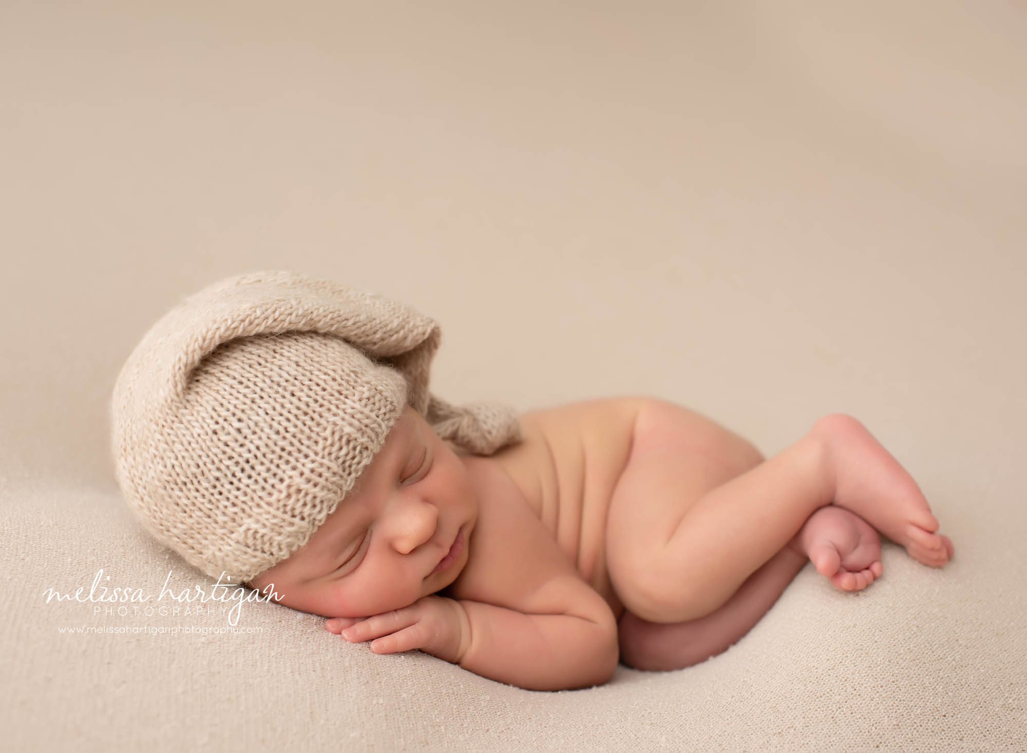 Baby boy posed side laying on tan colored blanket backdrop with knitted tan sleepy cap