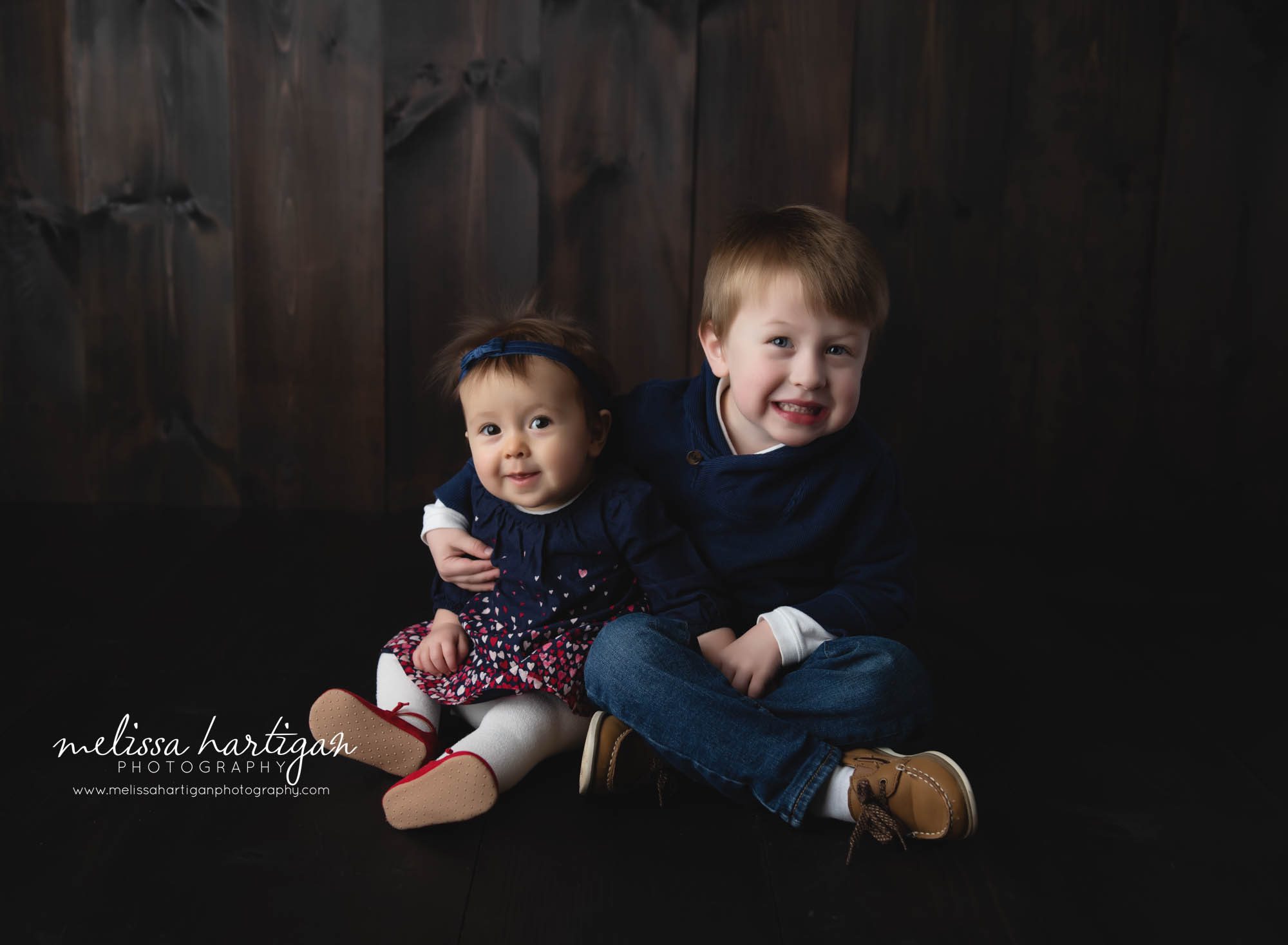 Baby girl sitting in studio with toddler big brother sitting beside her with his arm around her