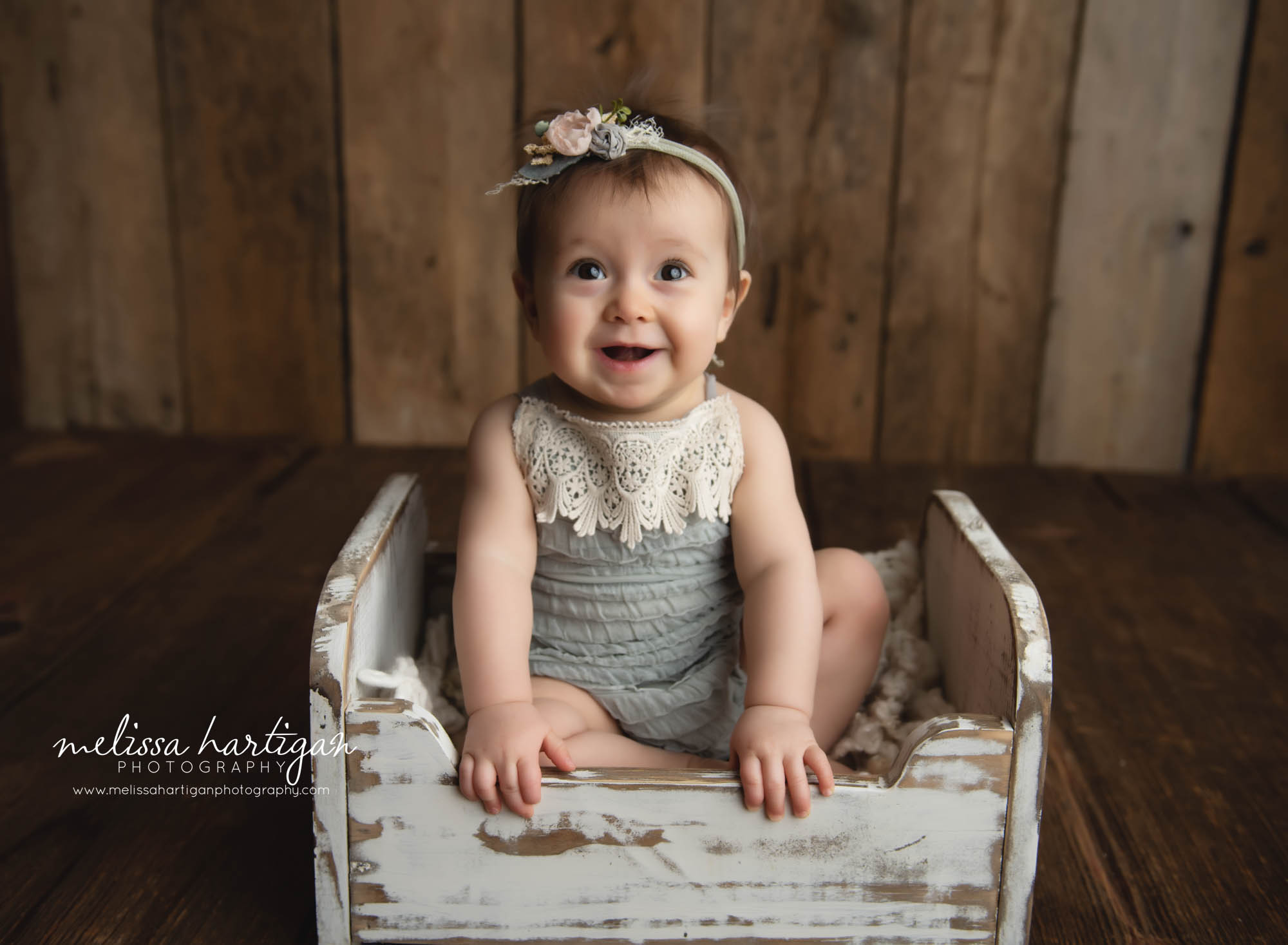 Baby girl sitting in vintage cream wooden cradle bed prop wearing light blue romper with cream lace