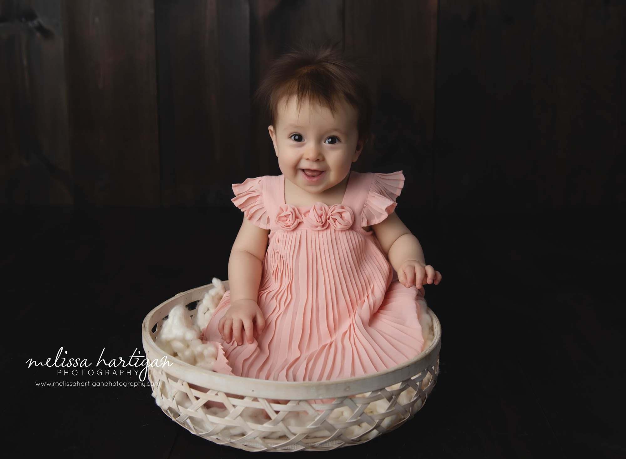 Baby girl sitting in cream wooden basket wearing pink dress with 3 rosebud flowers