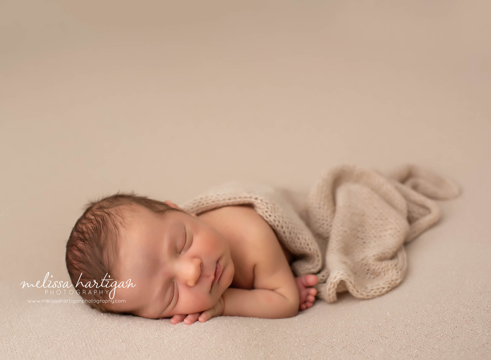 Baby boy posed on tummy with hand under cheek and toes peeking out with wrap lightly laying over him on tan colored backdrop newborn baby boy sleeping newborn photography session CT newborn photographers studio