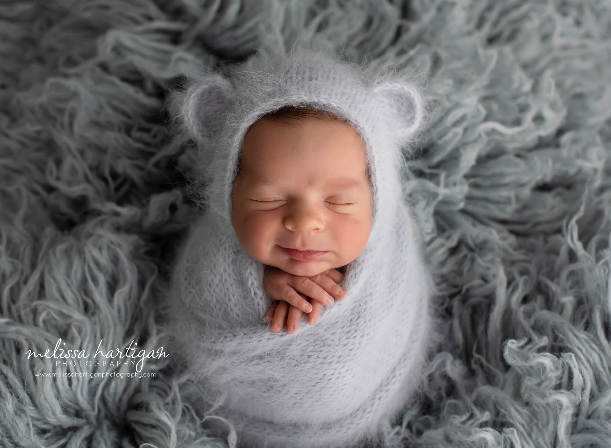 Newborn boy wrapped in light gray knitted wrap and matching knitted bear bonnet on gray flokati