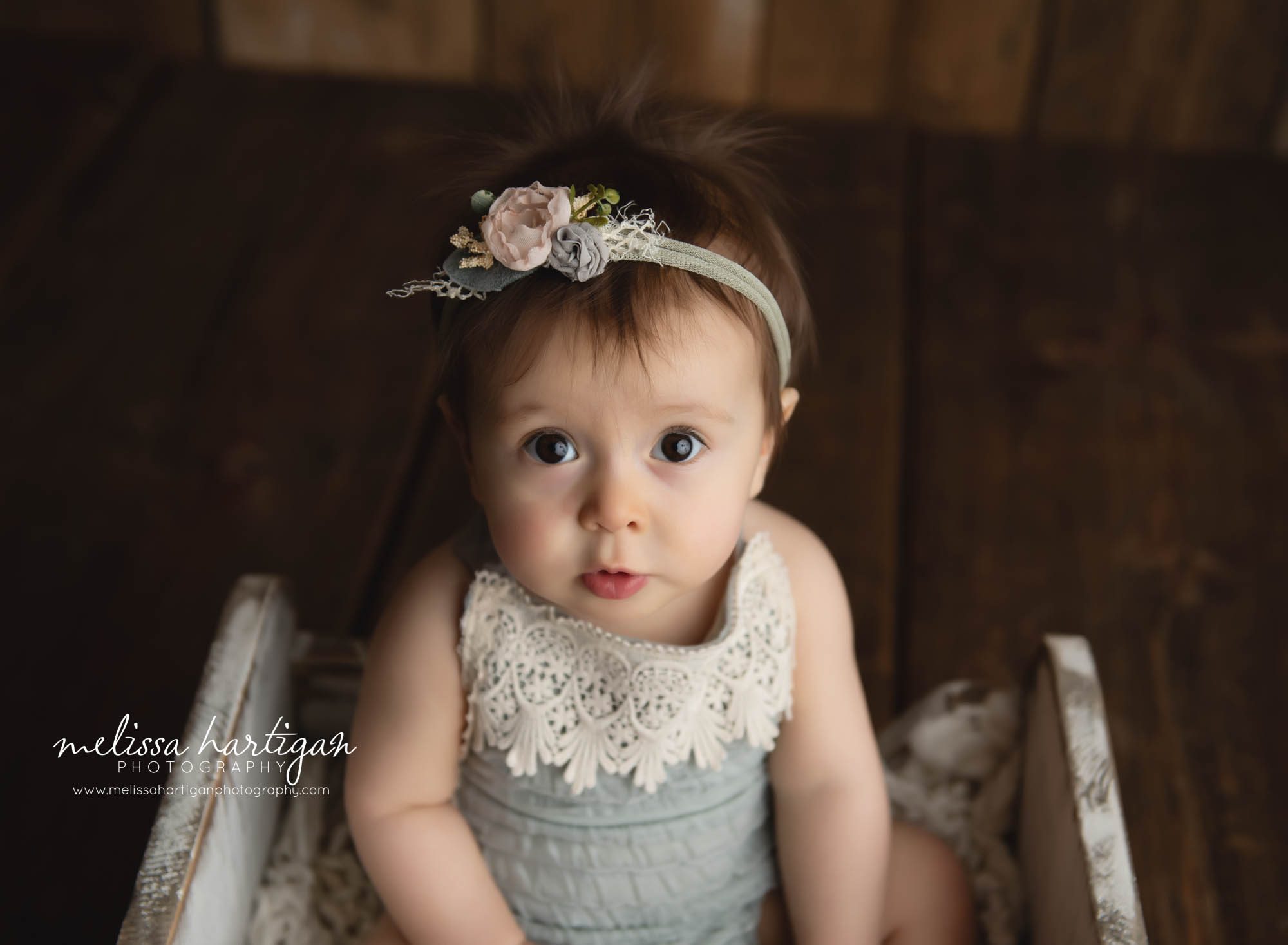 Baby girl sitting in wooden prop with blue romper with lace trim and floral headband