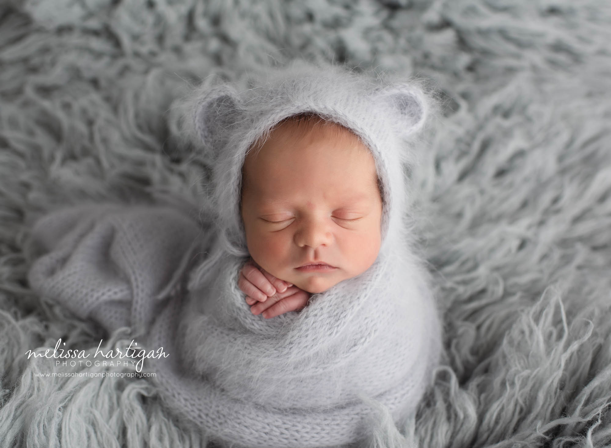 Baby boy wrapped in knitted light grey wrap and bear bonnet on grey flokati