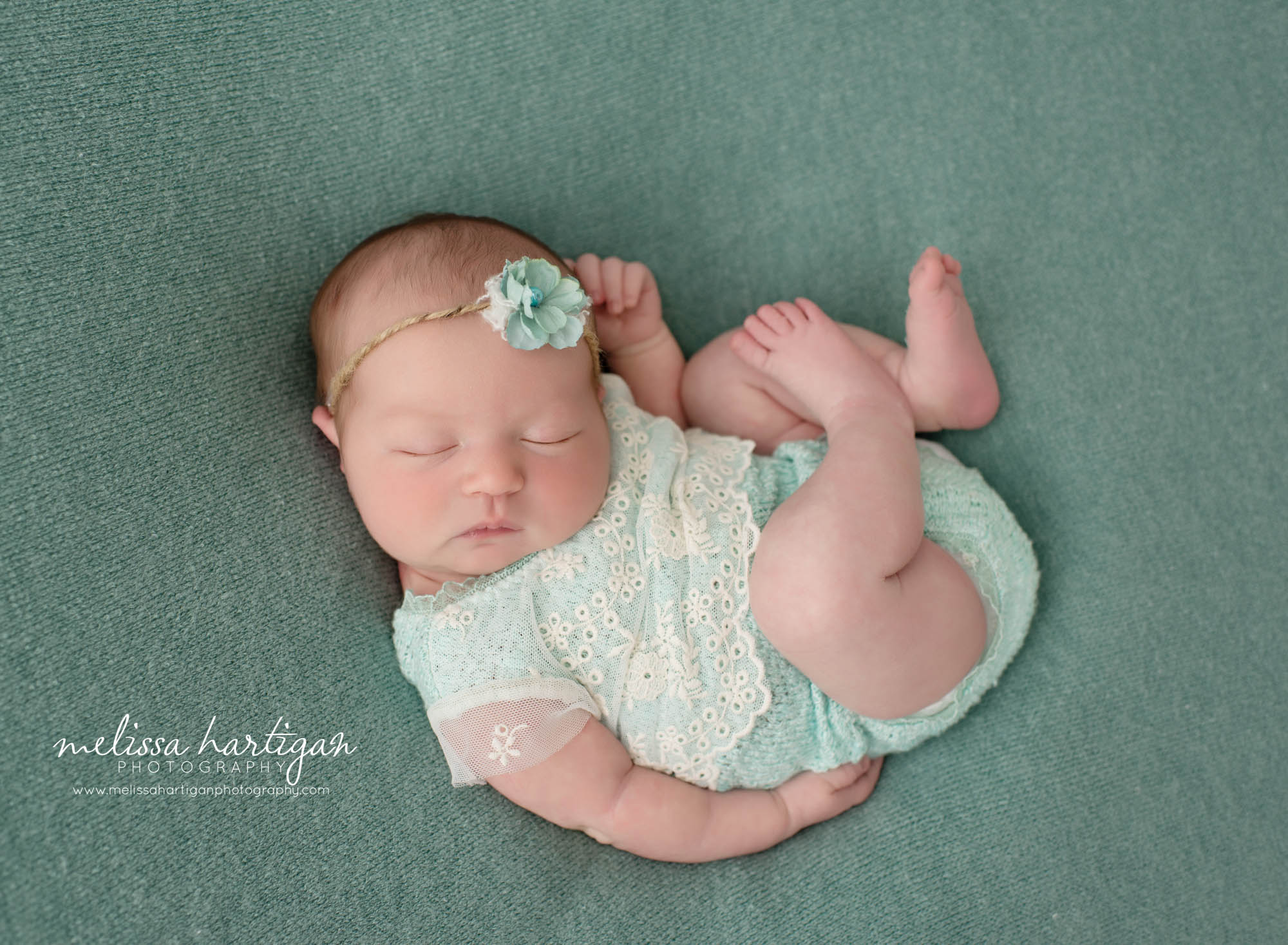 Baby girl posed on back in relaxed pose with teal colored eyelet romper and flower tieback