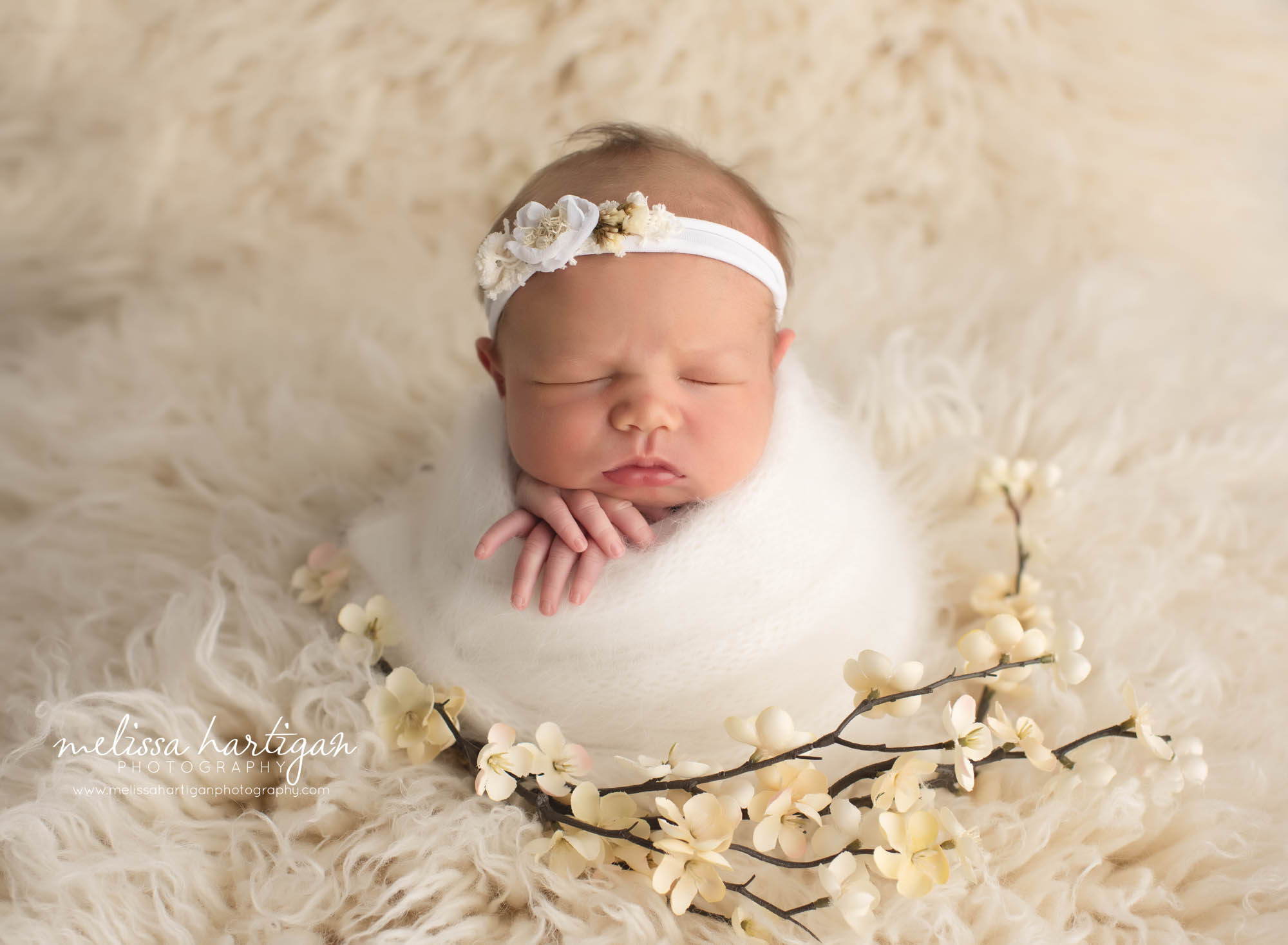 Baby girl wrapped in white wrap on flokati with floral headband and branch of flowers