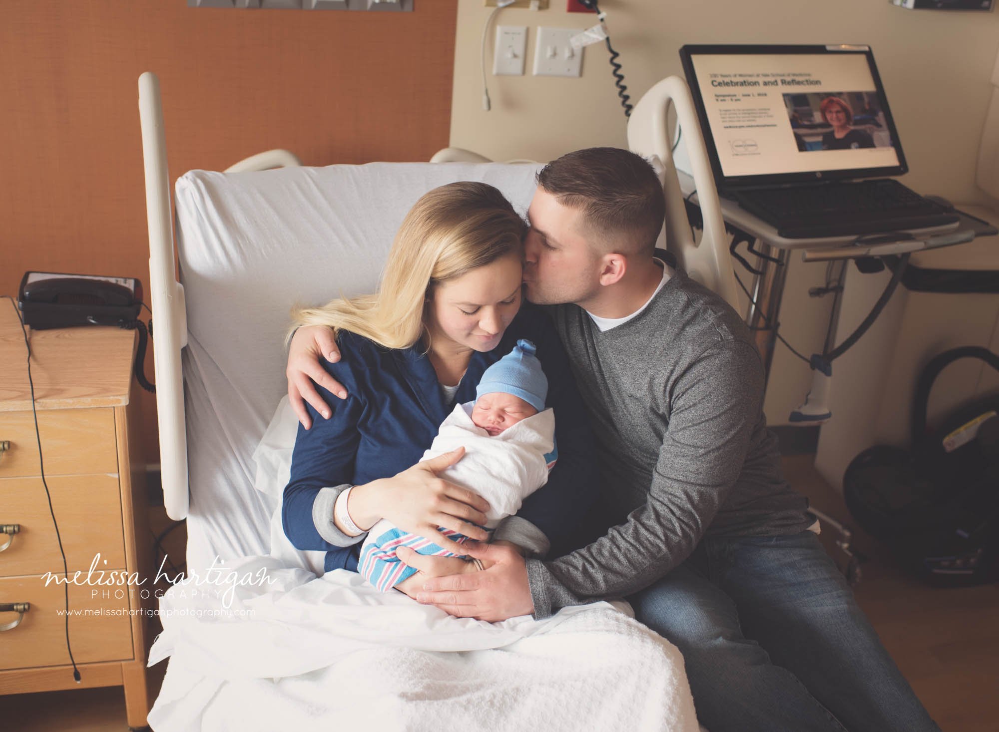 mom holding newborn baby boy sitting on hopsital bed with dad sitting next to mom and giving her kiss on the side of the head