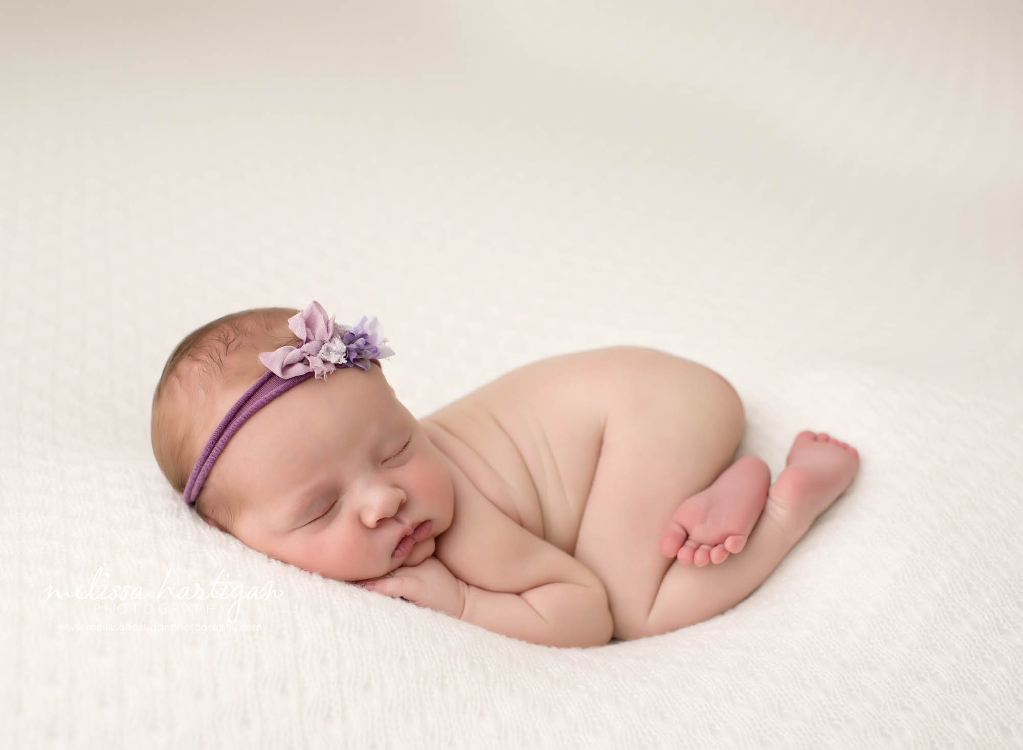 baby girl posed bum up pose on cream colored backdrop with purple flower headband