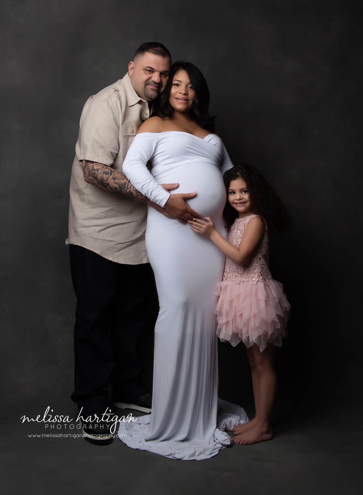 maternity family photo during in studio portrait session mom dad and daughter standing holding moms baby belly