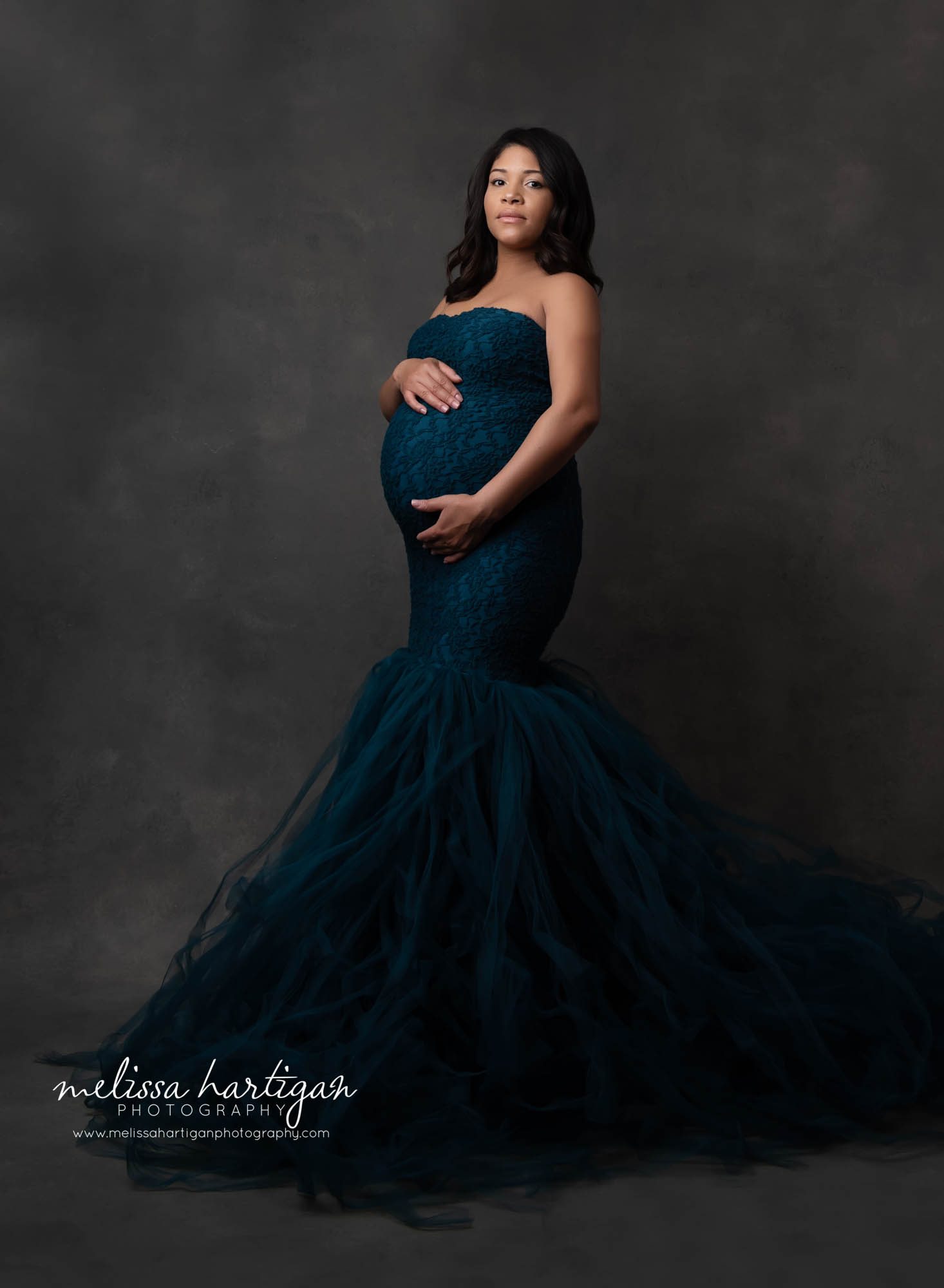 Expectant mom standing in photography studio during maternity session mom wearing teal colored slim fitting mermaid style maternity gown with lace overlay and tulle bottom skirt