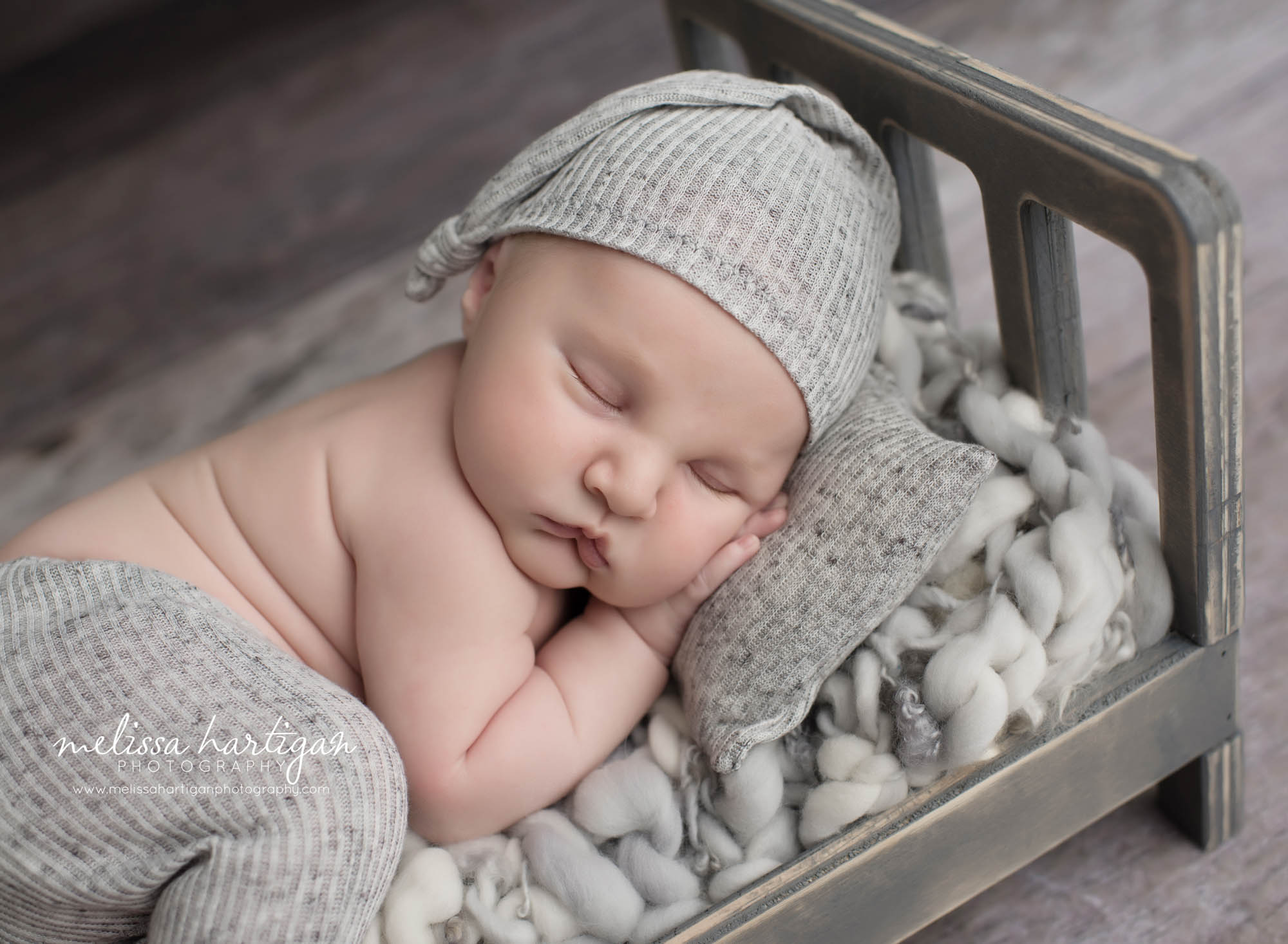 baby boy posed on wooden prop bed with gray pants hat and newborn pillow sleeping
