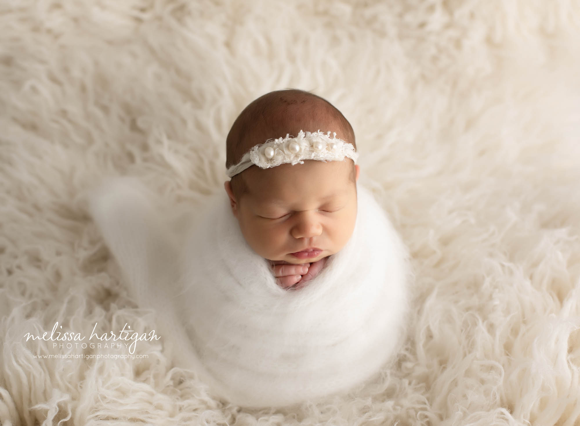 baby girl wrapped in knitted wrap with cream colored headband with pearls posed on flokati rug newborn photography session