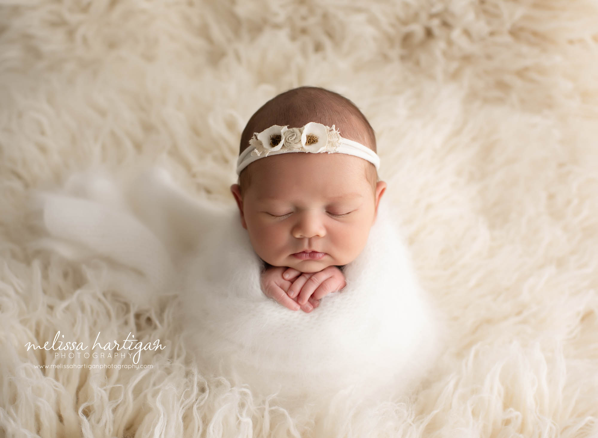 baby girl wrapped in white knitted wrap posed on cream flokati with flower headband