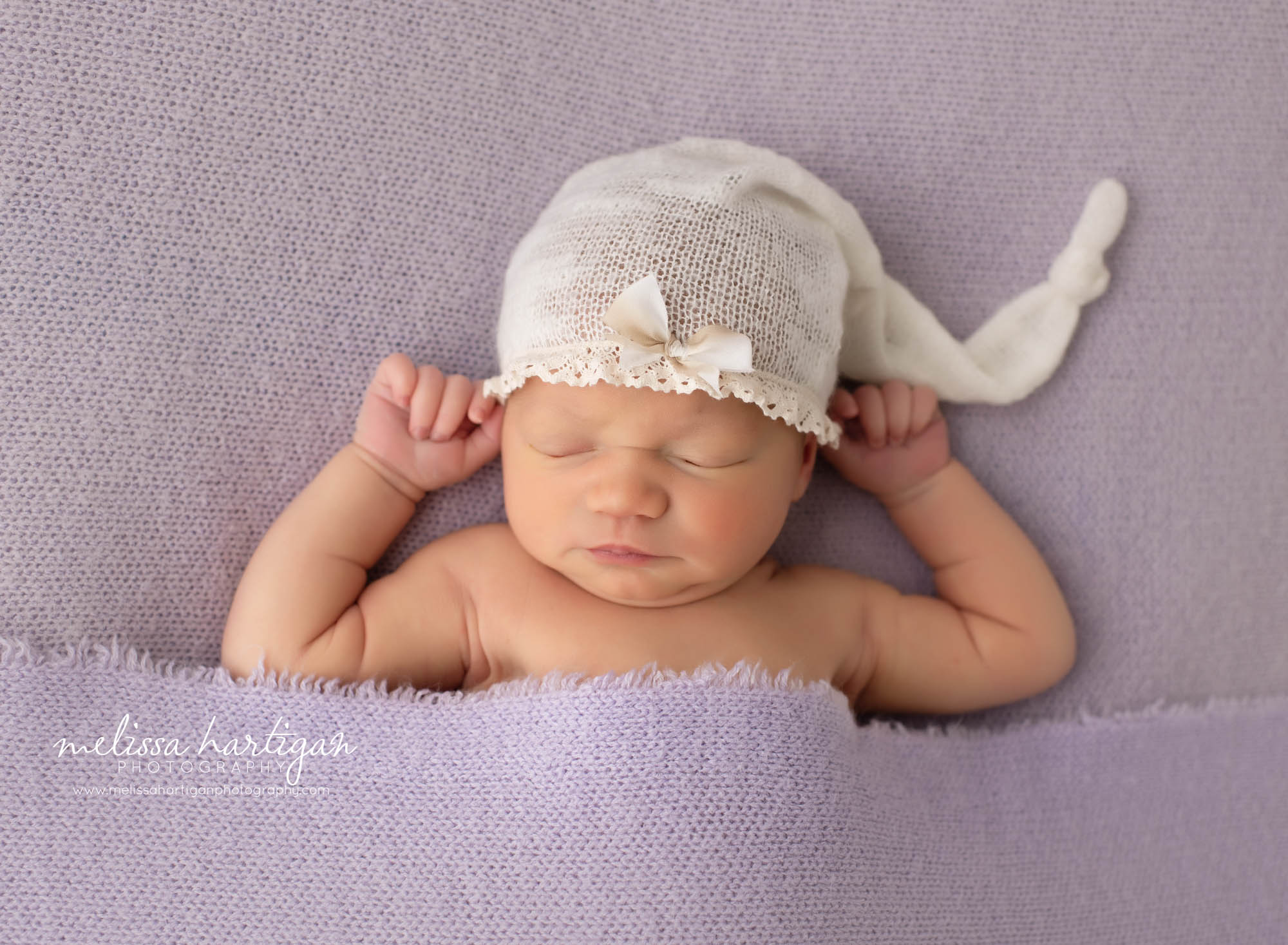 baby girl laying on back with purple blanket and cream sleepy cap