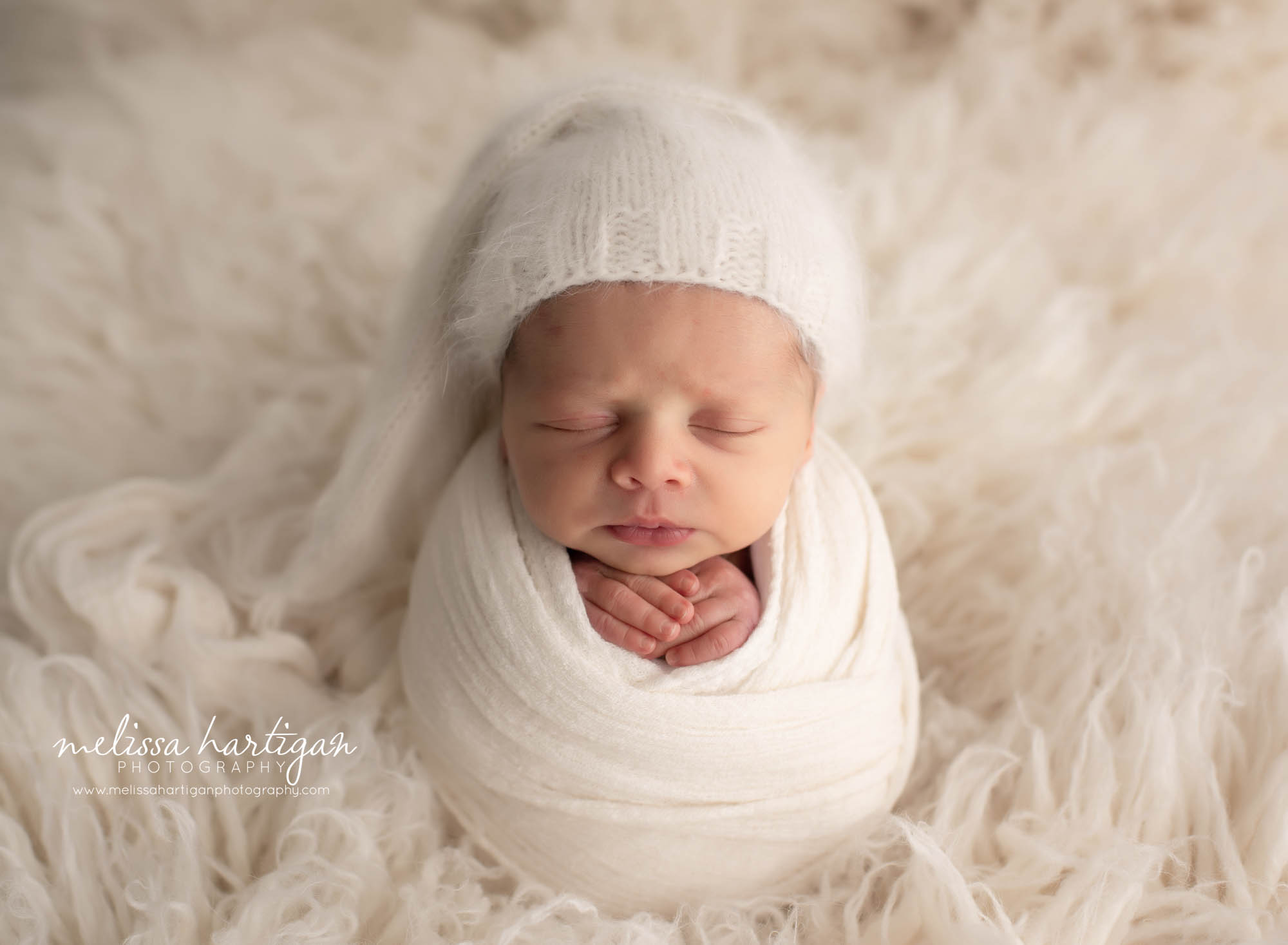 baby wrapped in cream colored wrap and wearing knitted sleepy cap on cream flokati rug
