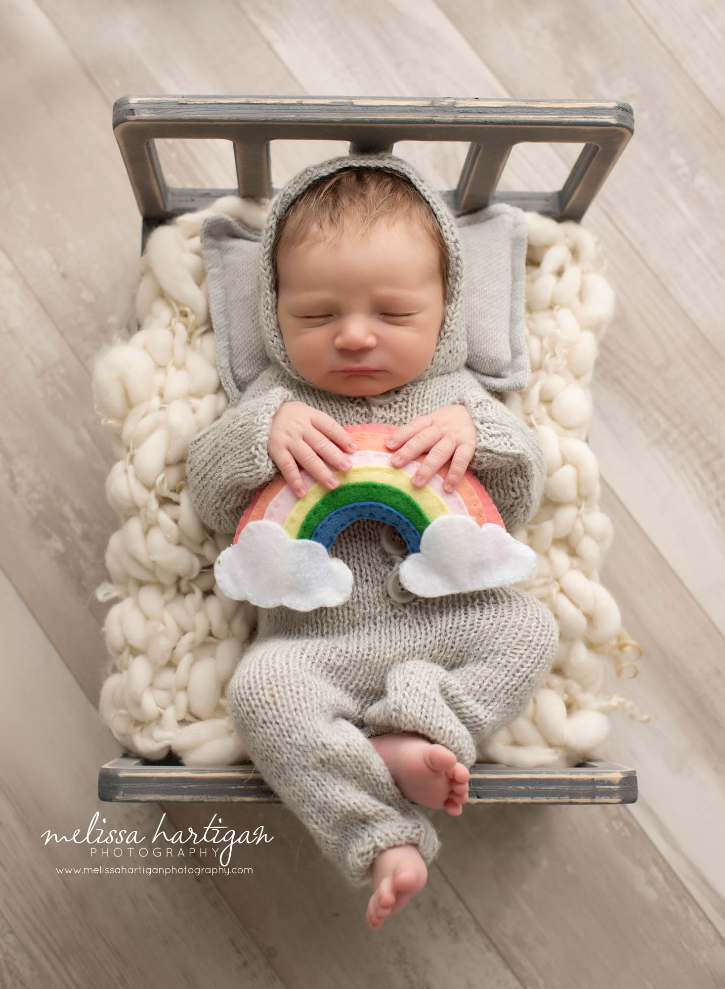 Newborn baby boy posed on wooden bed laying on back wearing grey outfit holding rainbow prop