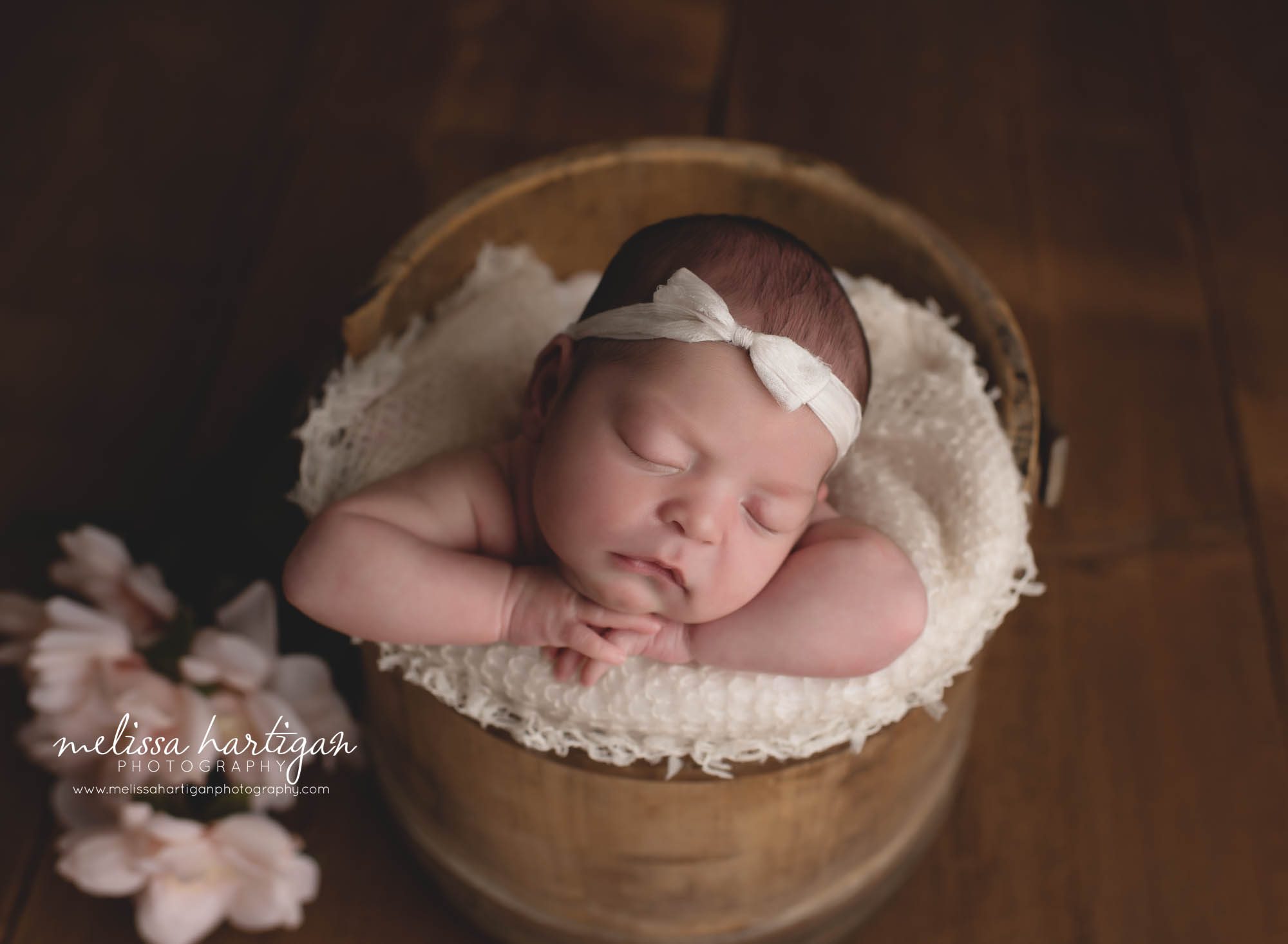 newborn baby girl posed in wooden bucket with head on hands wearing bow headband