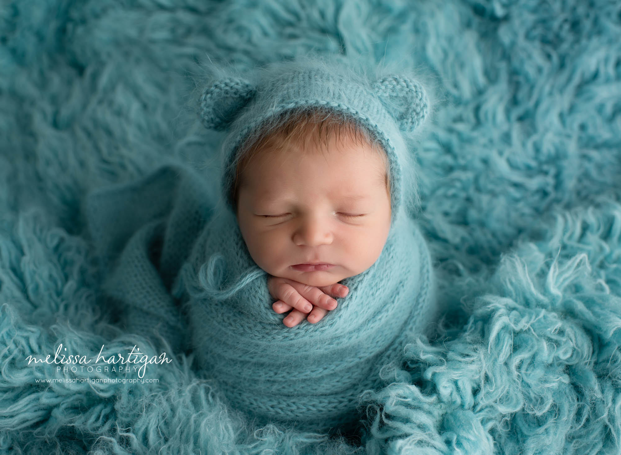 Baby boy wrapped in teal knitted wrap and matching bear bonnet on tal colored flokati