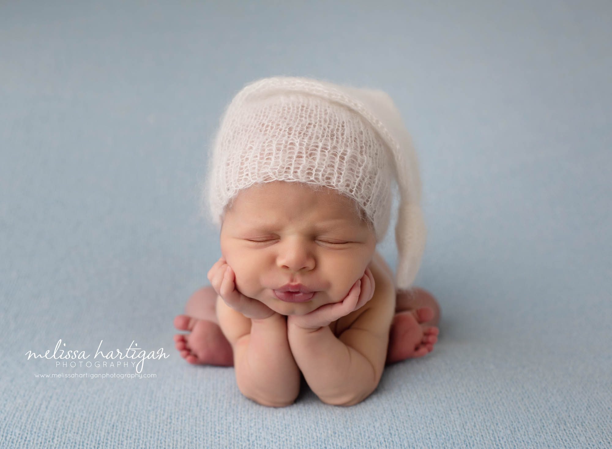baby boy posed froggy pose on light blue backdrop wearing knitted sleepy cap