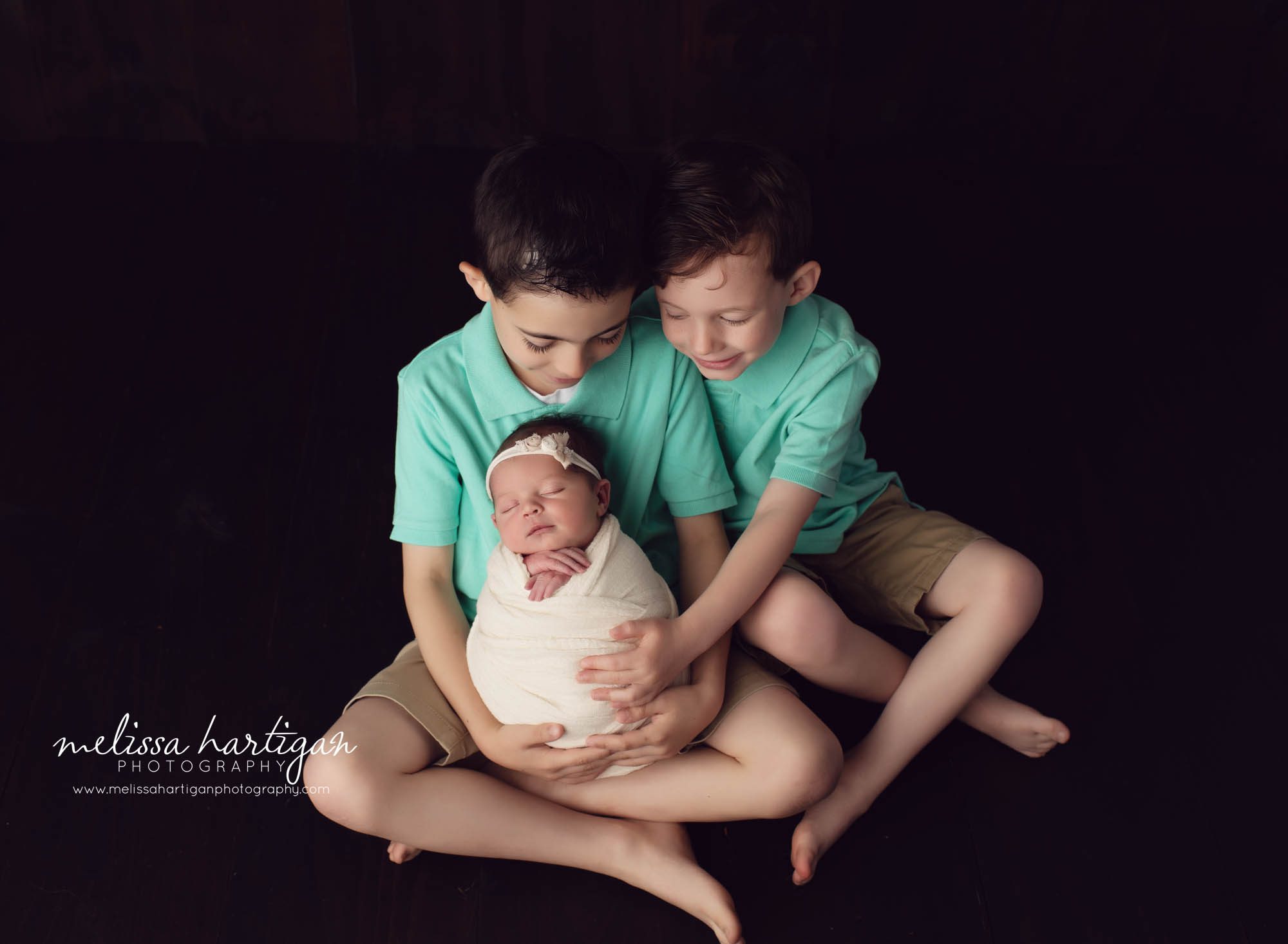 two big brothers holding baby sister wrapped in wrap wearing flower headband