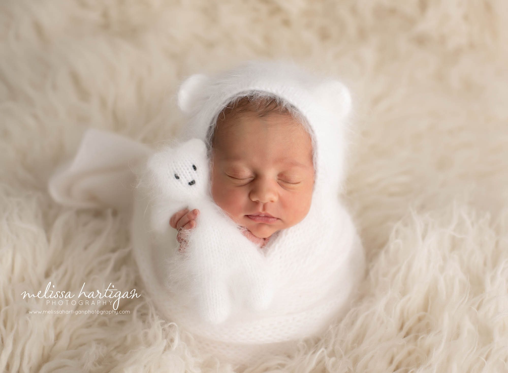 newborn baby wrapped in white wrap with matching bear bonnet holding miniature knitted teddy bear