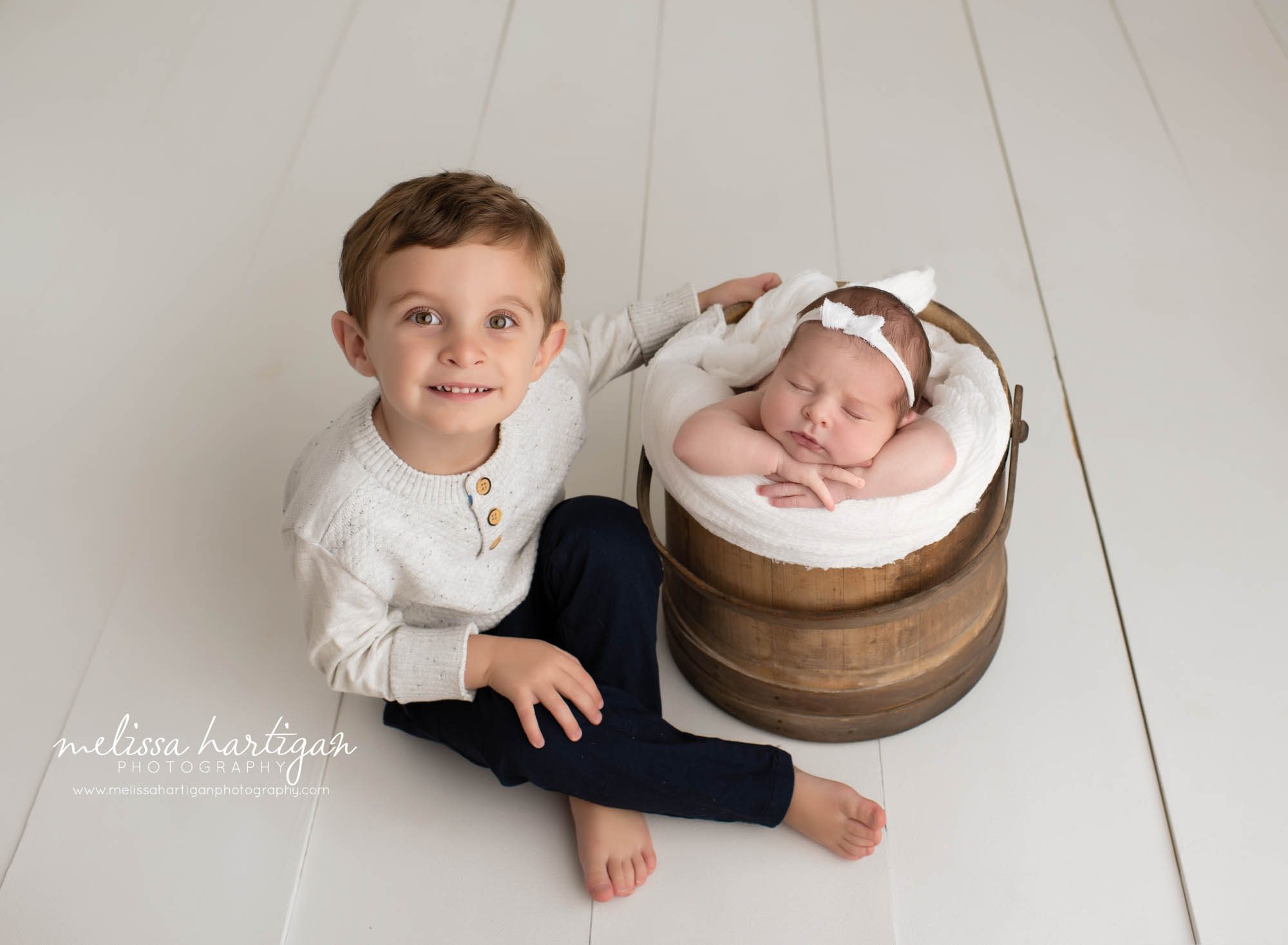big brother sitting beside newborn baby sister posed in wooden rustic bucket with white layer and white bow headband