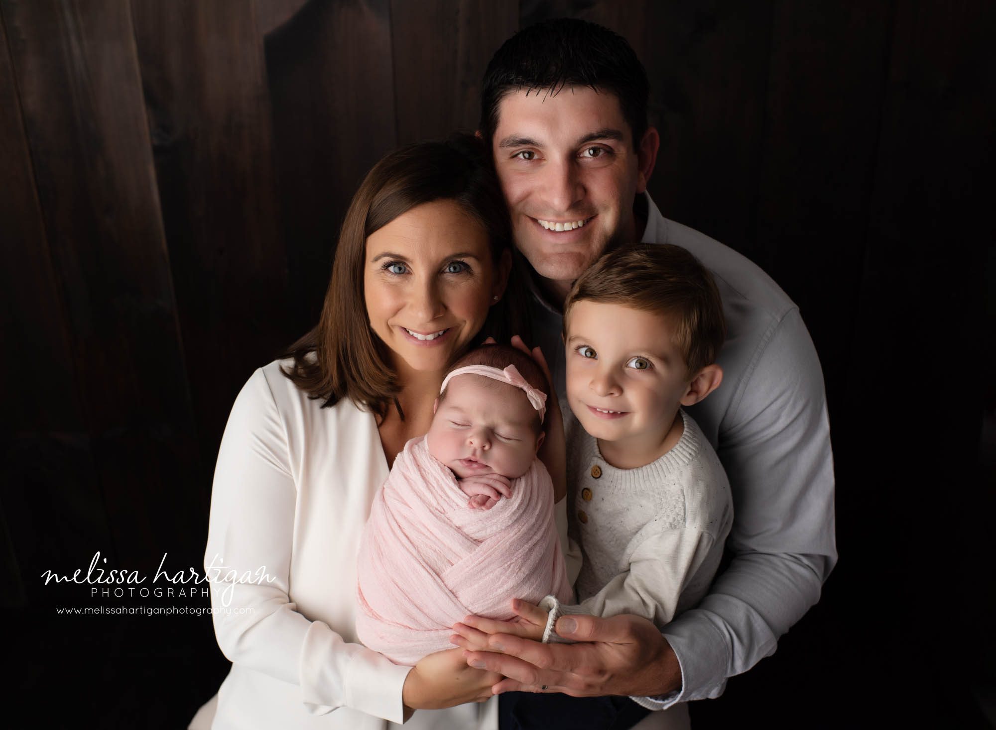 mom dad and big brother holding newborn baby girl wrapped in light pink wrap studio photography session