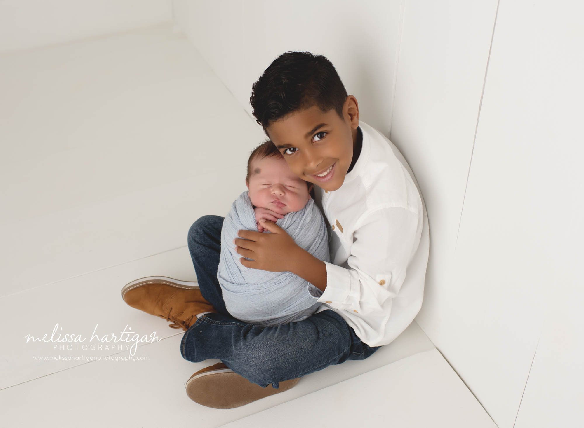 older brother sitting on wooden board floor holding wrapped newborn baby brother and smiling at camera for picture