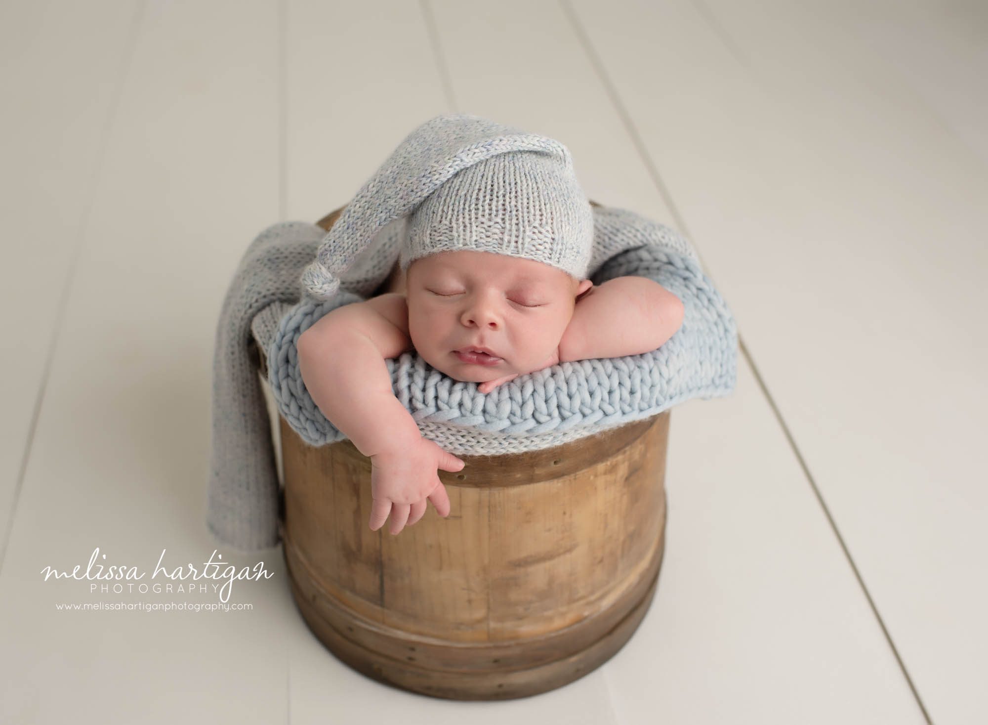 Baby boy posed in wooden prop bucket with blue toned knitted layer blanket and knitted sleepy cap