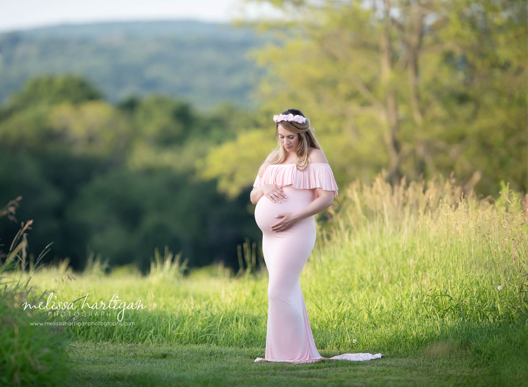 Pregnant mom standing in field looking down at baby bump wearing pink form fitting maternity dress with ruffle top and pink floral crown maternity Photographer CT